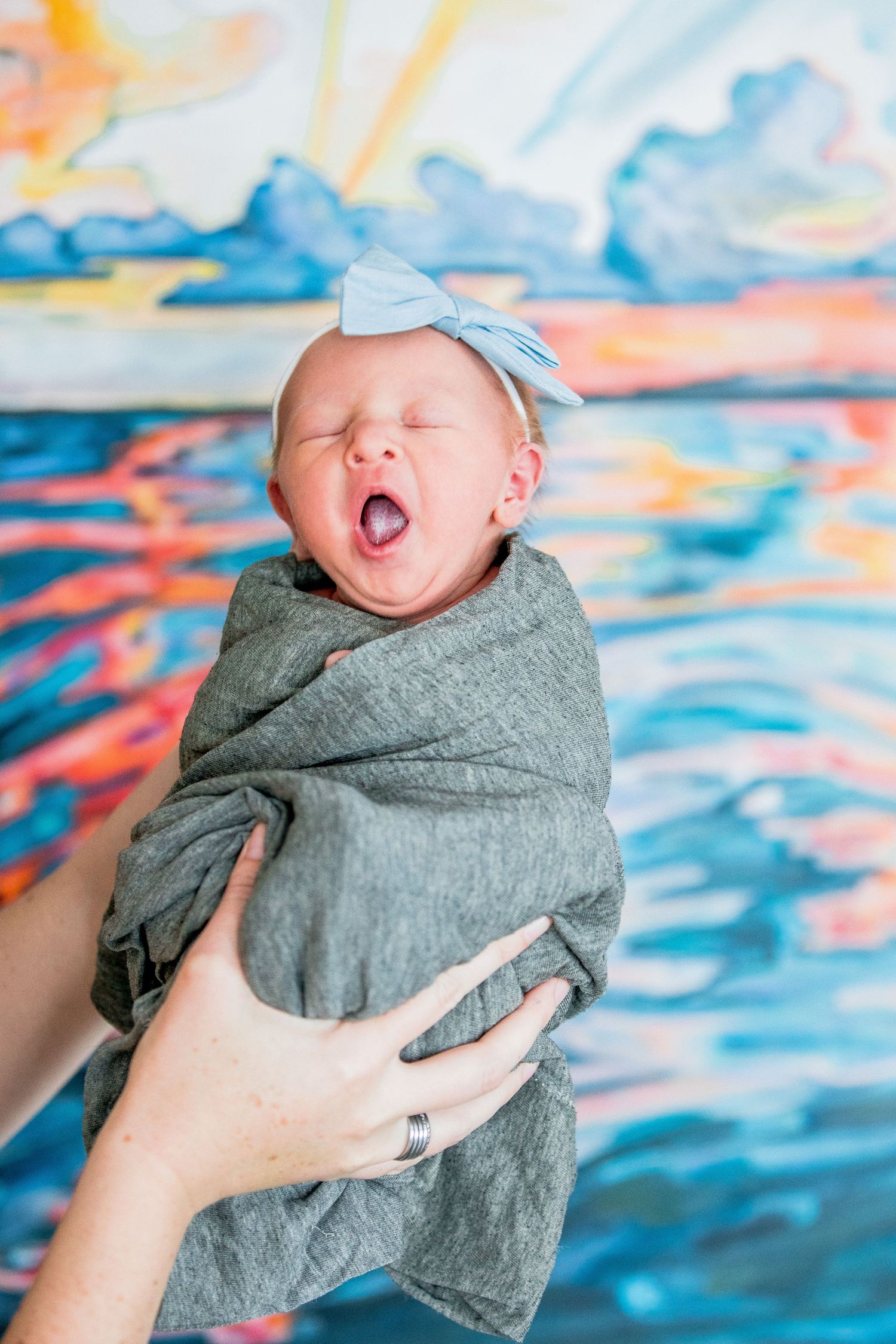 Yawning newborn baby swaddled in gray wrap with blue bow headband held up against colorful painting.