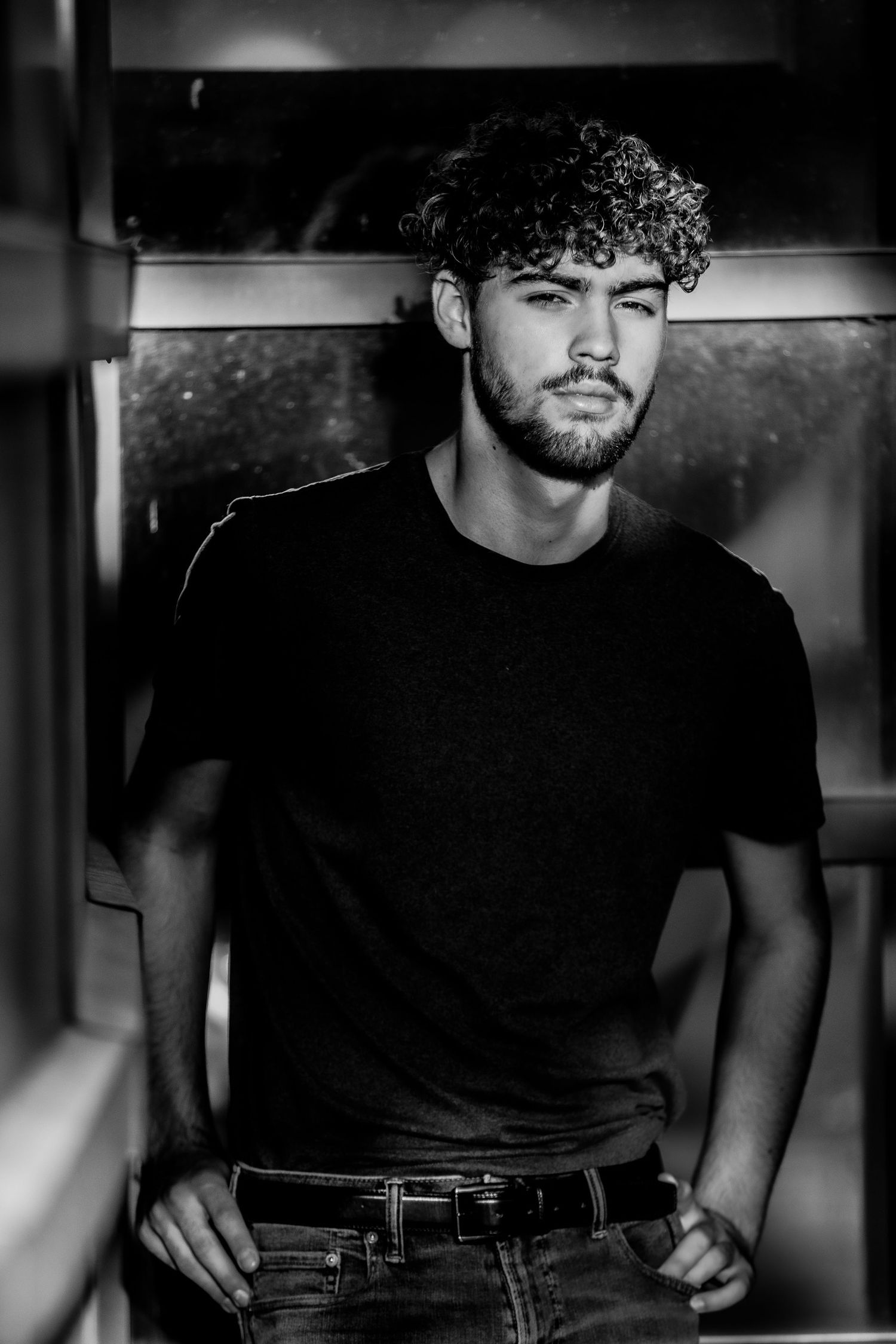 Black and white portrait of a young man with curly hair in a black t-shirt indoors.