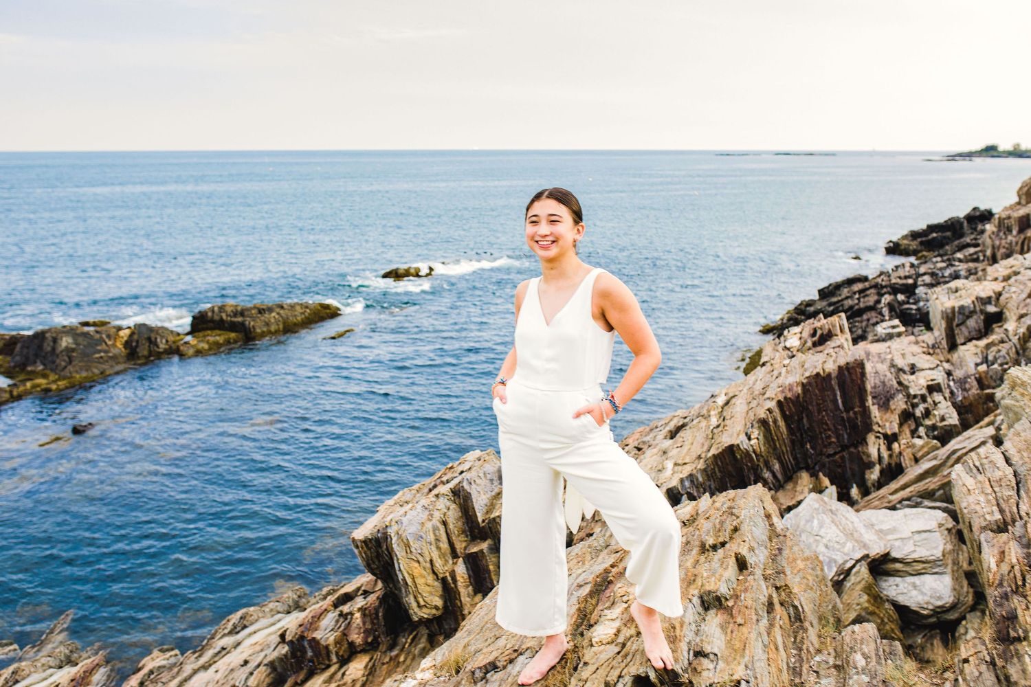 Smiling woman in white linen outfit posing on coastal rocks with bright blue ocean in the background.