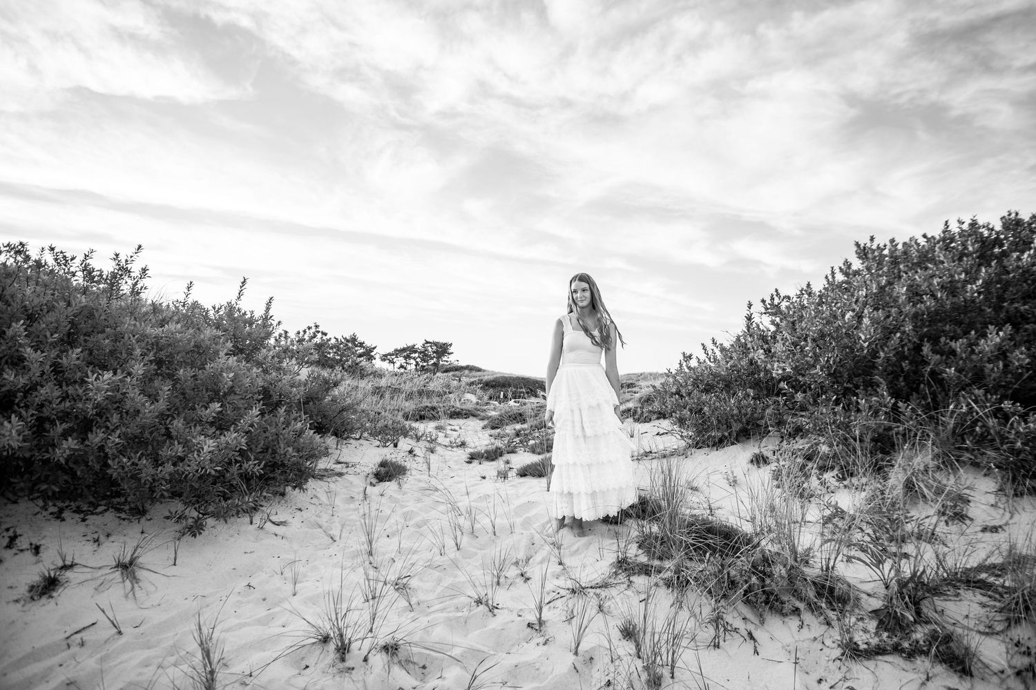 Woman in white dress standing among sand dunes and coastal shrubs in a serene black and white beach landscape.