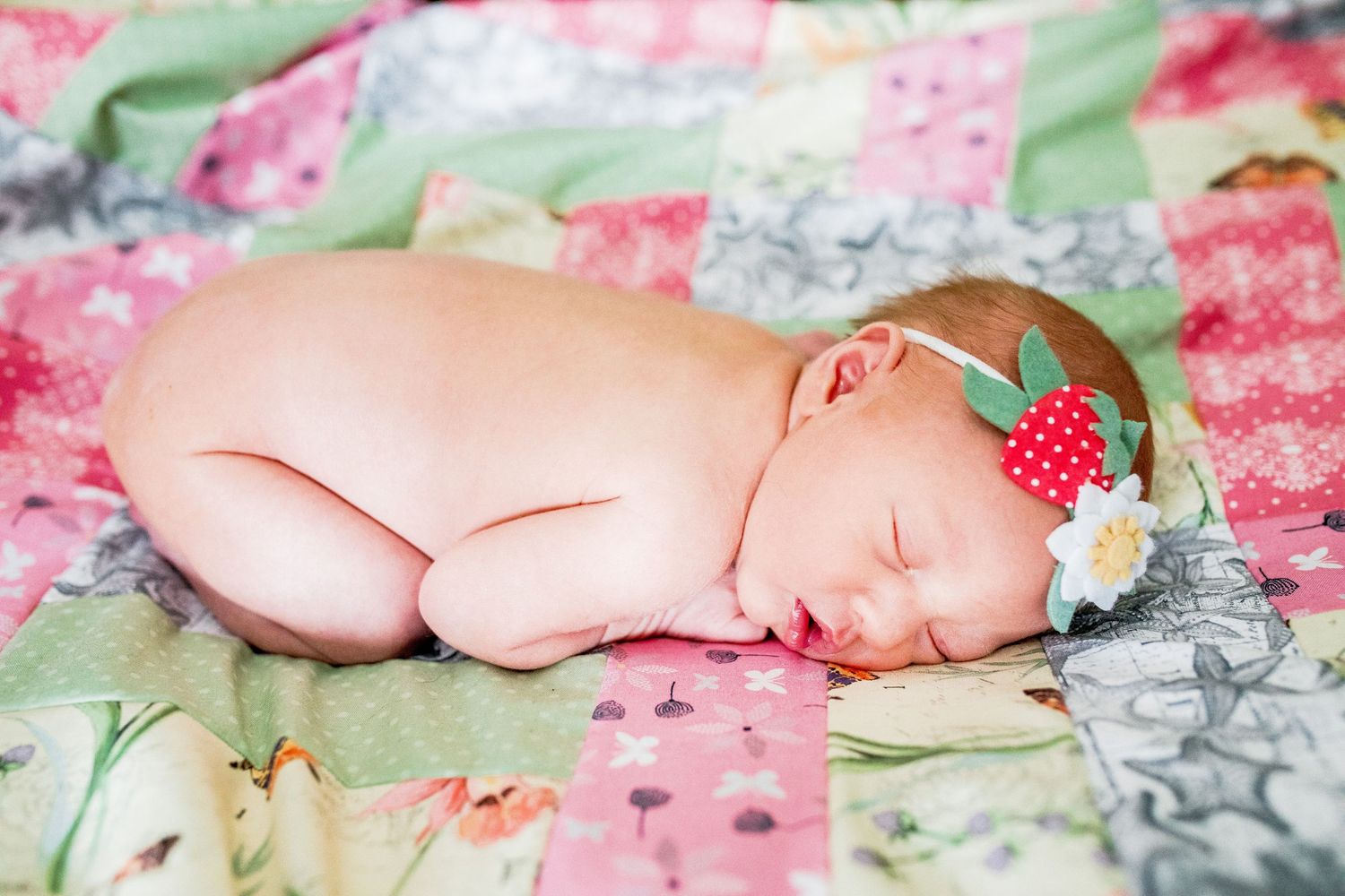 Sleeping newborn baby girl with floral headband curled up on colorful patchwork quilt.