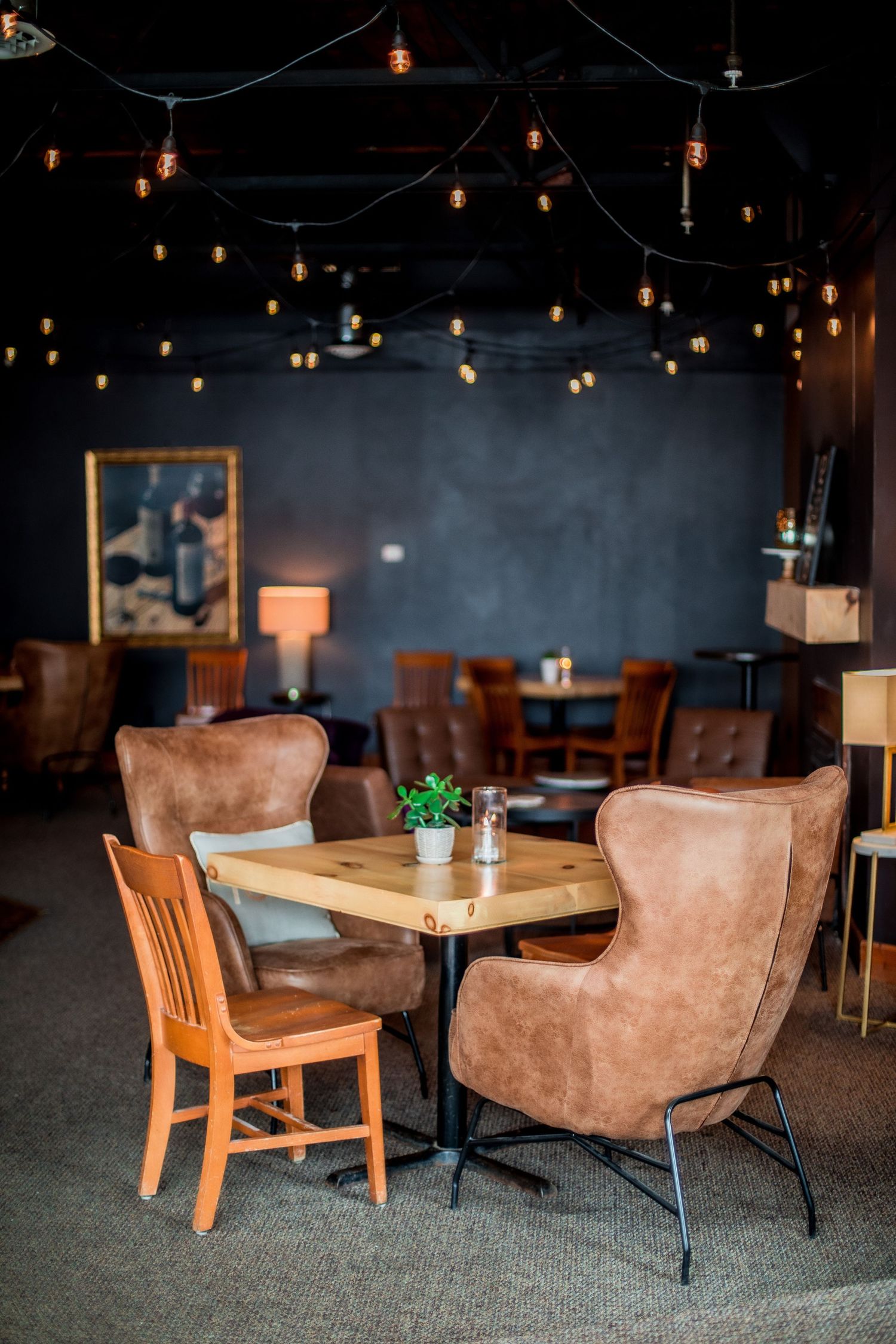 Cozy restaurant interior with brown leather chairs, wooden tables, Edison bulb string lights, and dark walls.