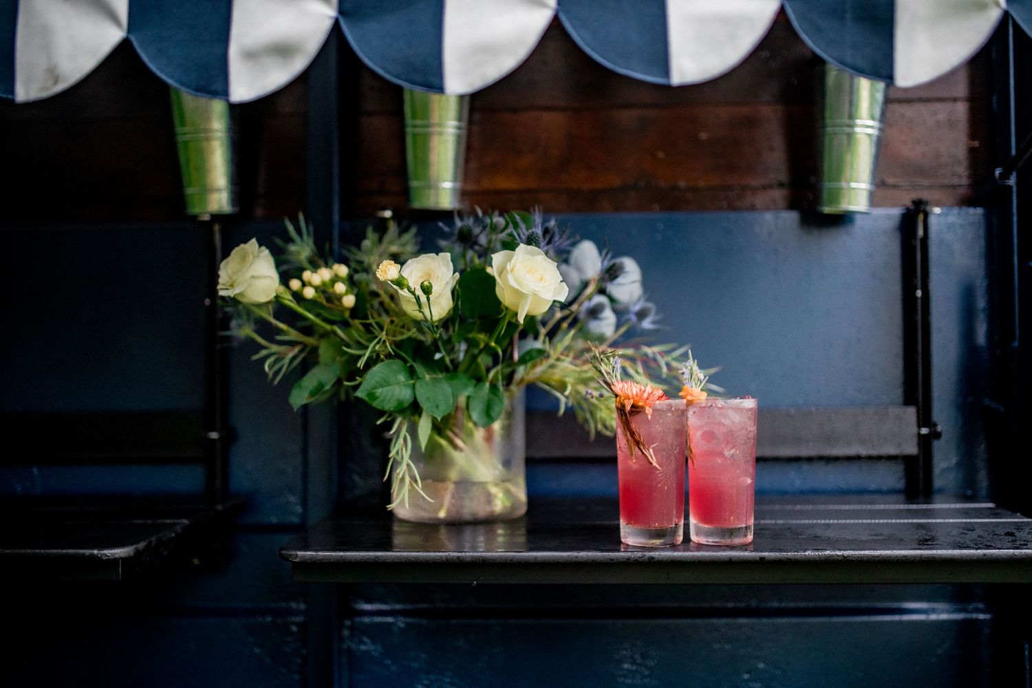 Pink cocktails and white rose bouquet displayed on dark wooden surface under striped awning.