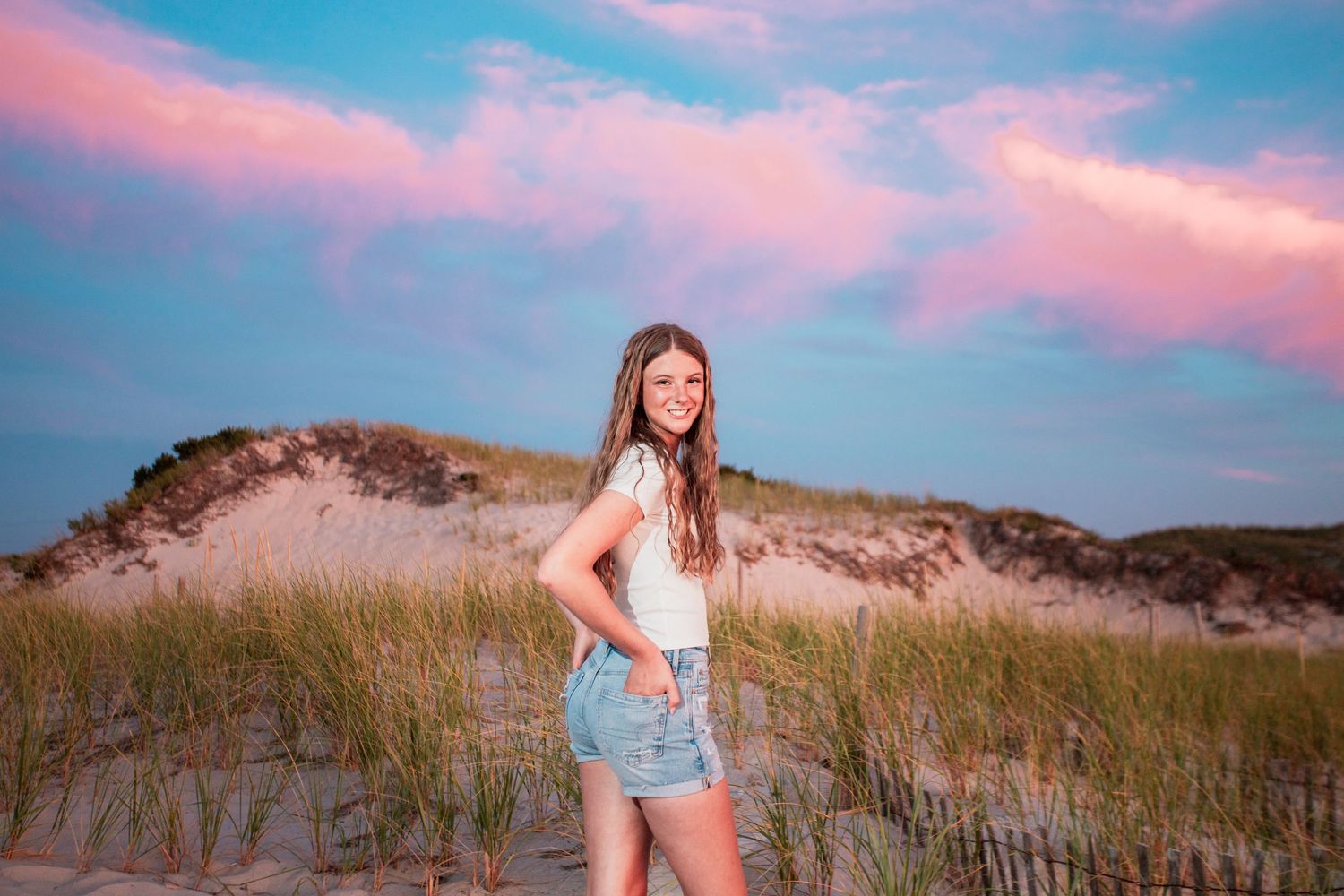 Young woman in denim shorts poses on sandy beach dunes under a stunning pink and blue sunset sky.