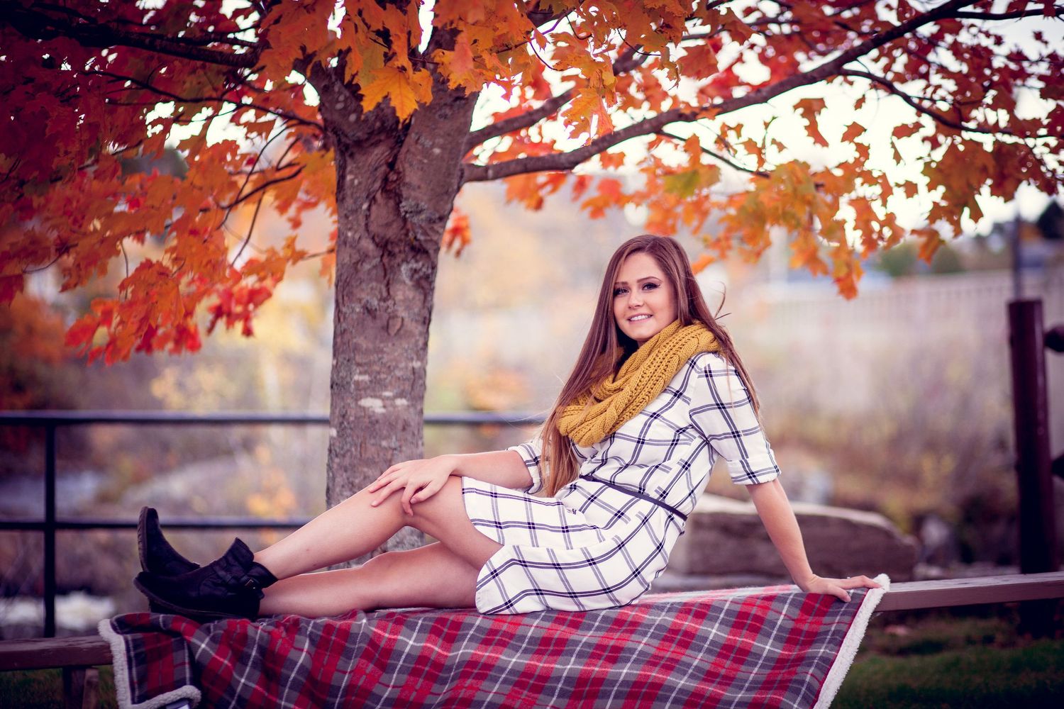 Young woman in plaid dress and yellow scarf sits on a bench beneath a vibrant red autumn maple tree.