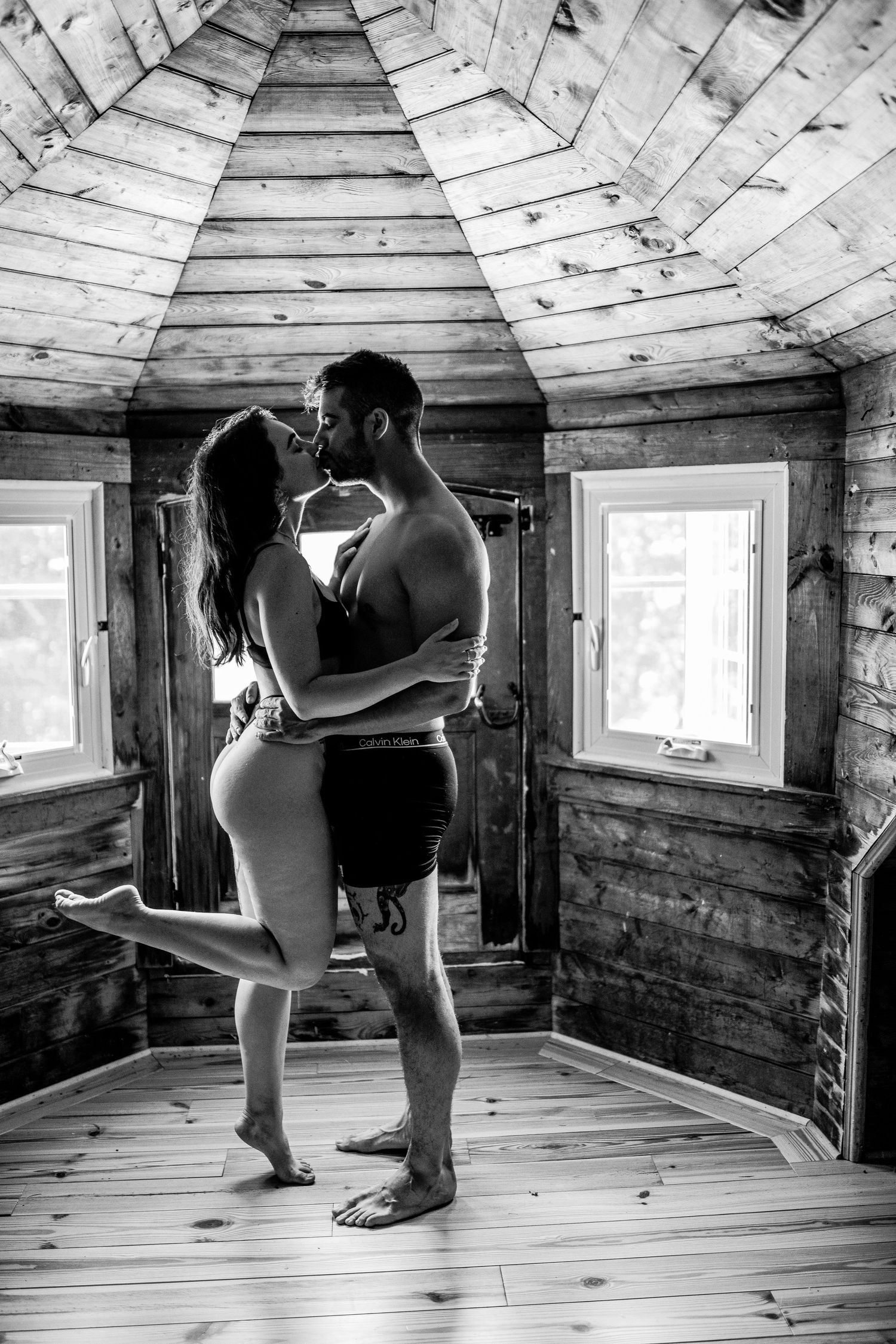 Black and white photo of a couple kissing and embracing inside a rustic wooden cabin room.