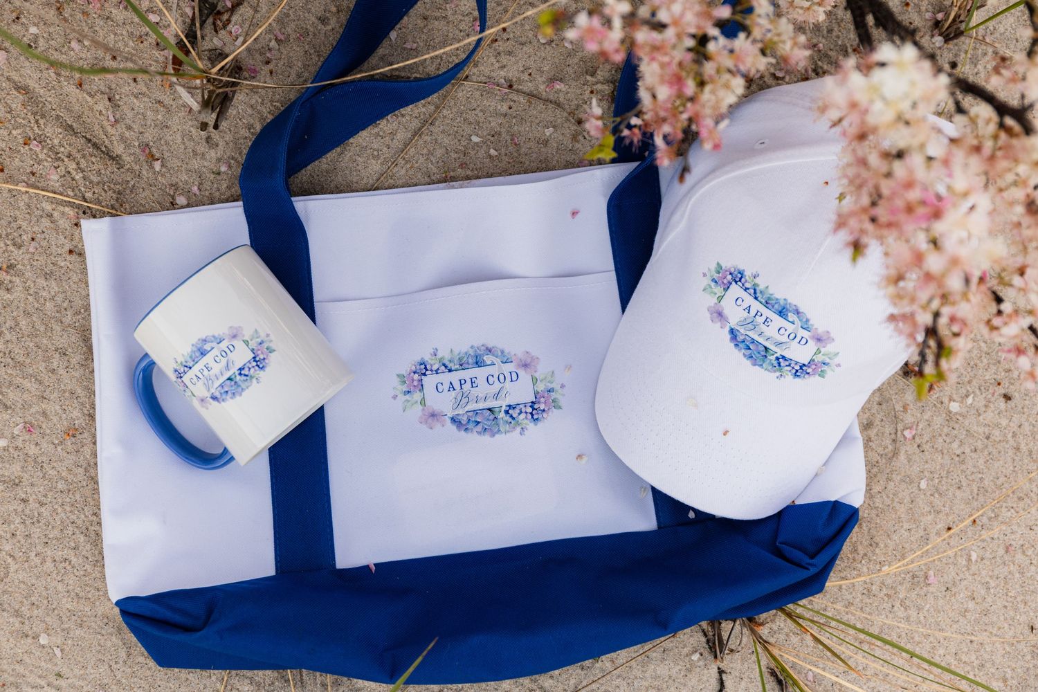 White tote bag, enamel mug, and baseball cap with matching Cape Cod logo branding on sandy surface.