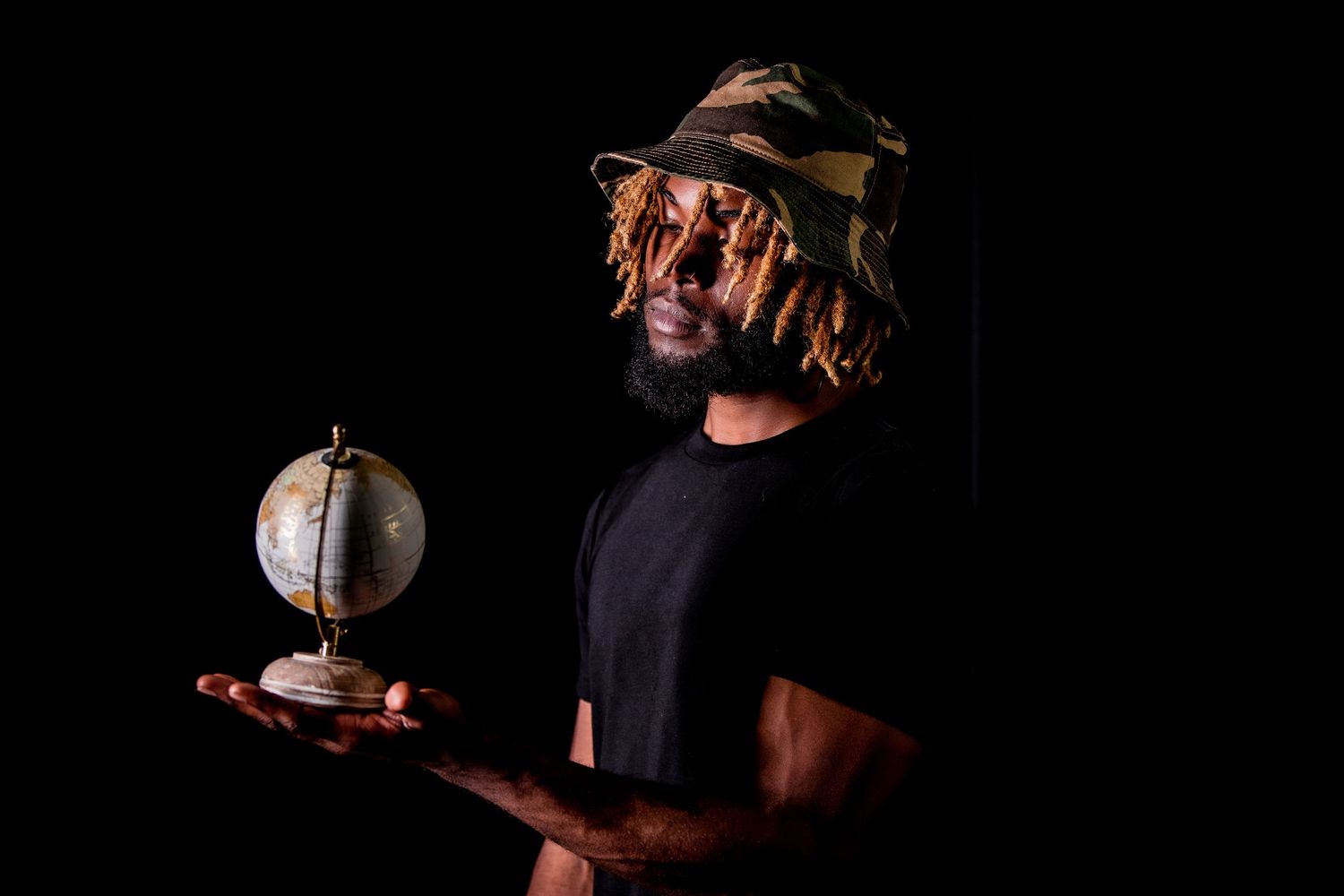 Man in camo bucket hat holding a small globe against a dark background.