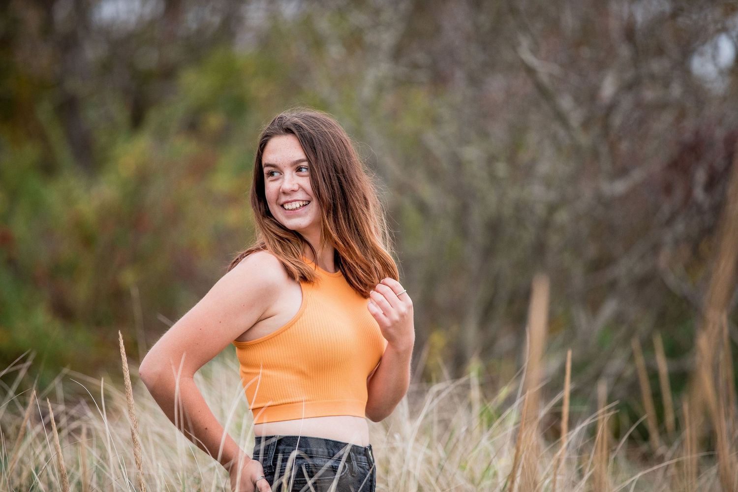 Happy teenage girl in orange crop top posing in tall grass with autumn foliage background.