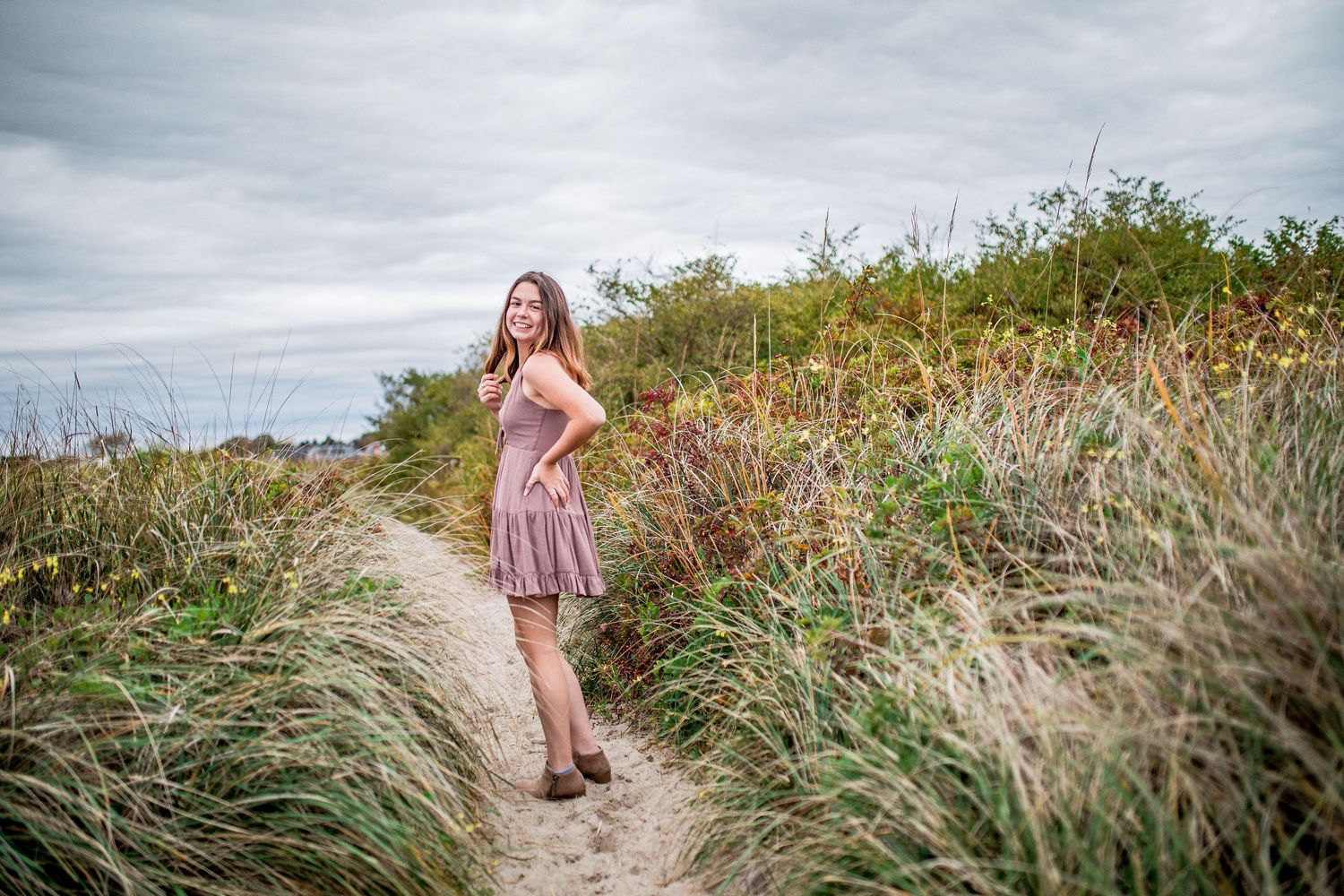 Young woman in pink dress smiling on a sandy coastal path surrounded by tall grass.