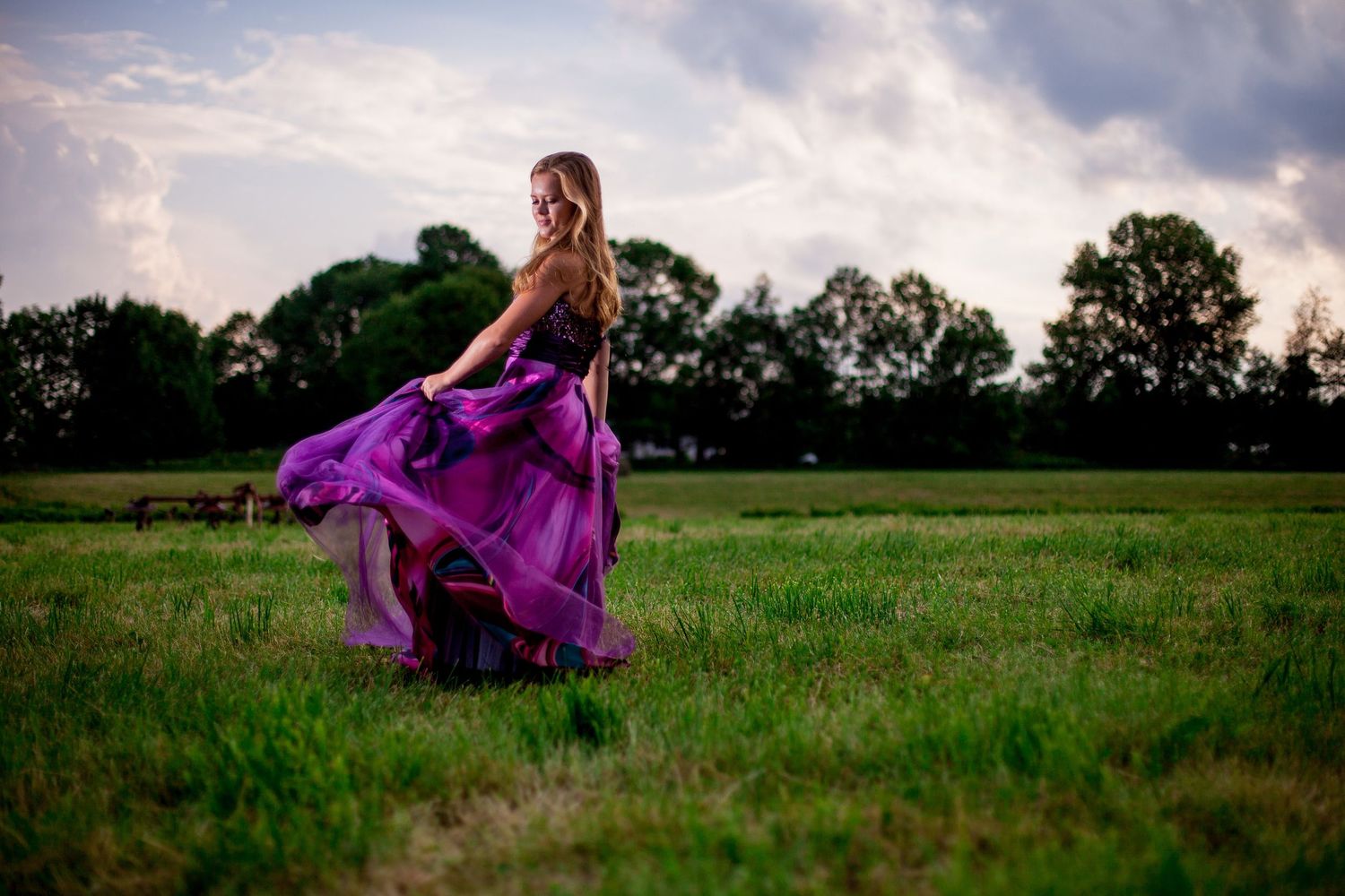 Young woman in a flowing purple dress walking through a lush green field at dusk.