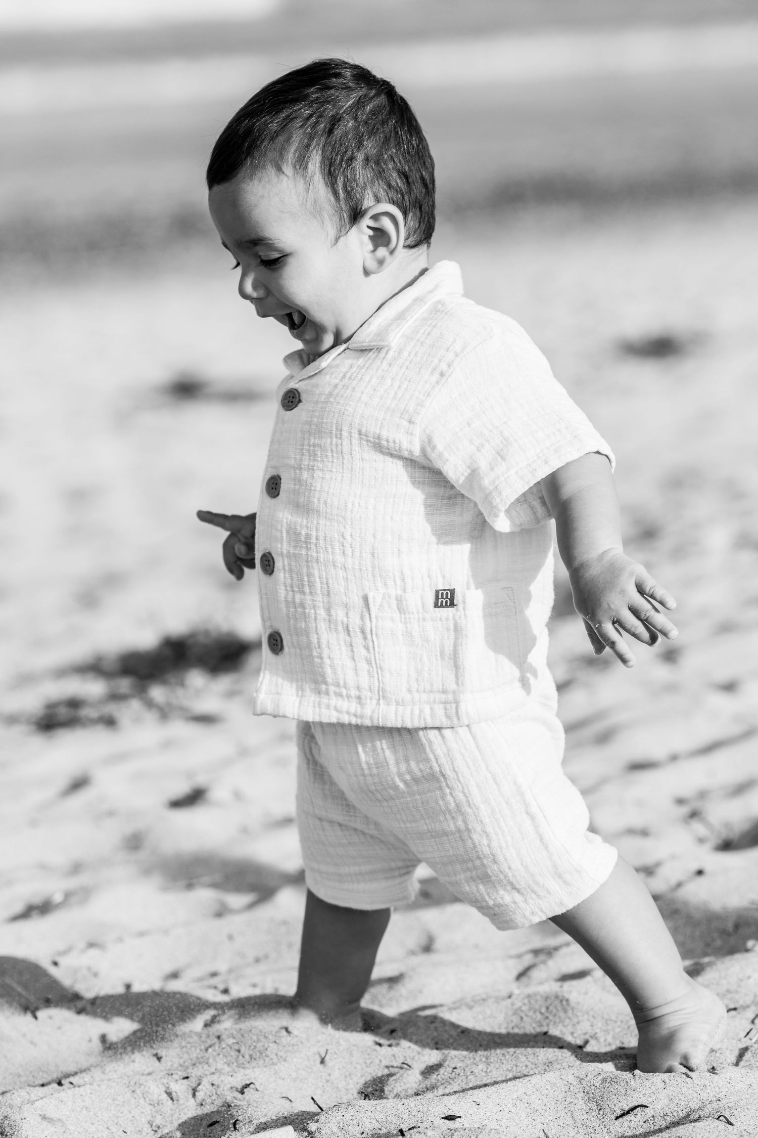 Black and white photo of a happy toddler boy walking on sandy beach wearing a linen shorts set.