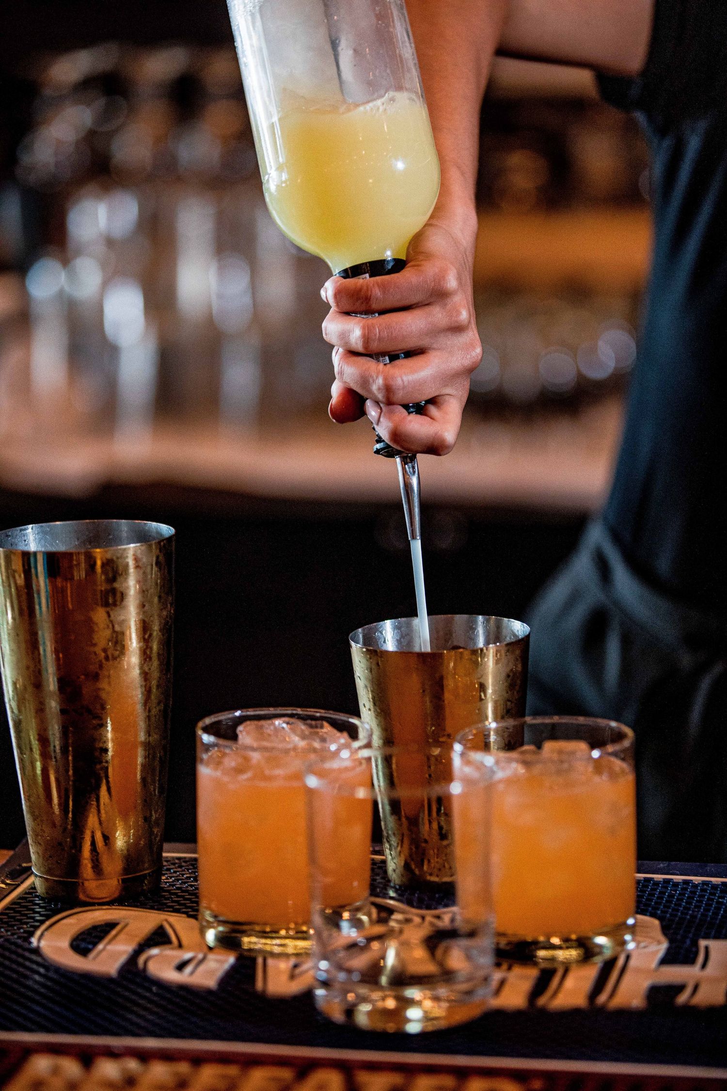 Bartender pouring fresh juice into gold cocktail shakers and glasses filled with ice at a bar.