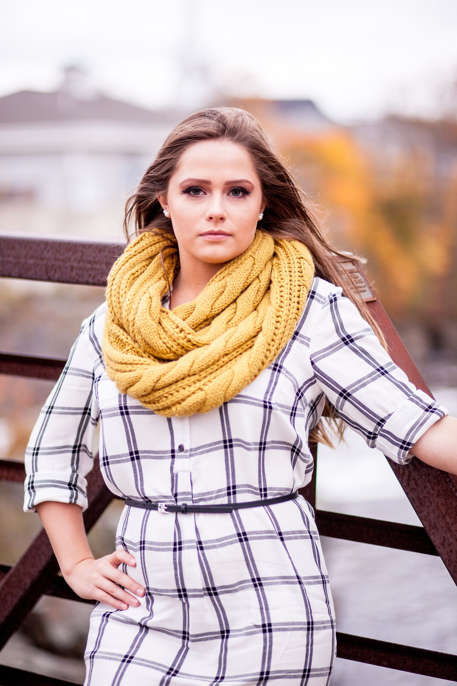 Young brunette woman in plaid dress and yellow infinity scarf leans against metal railing with autumn trees behind her.