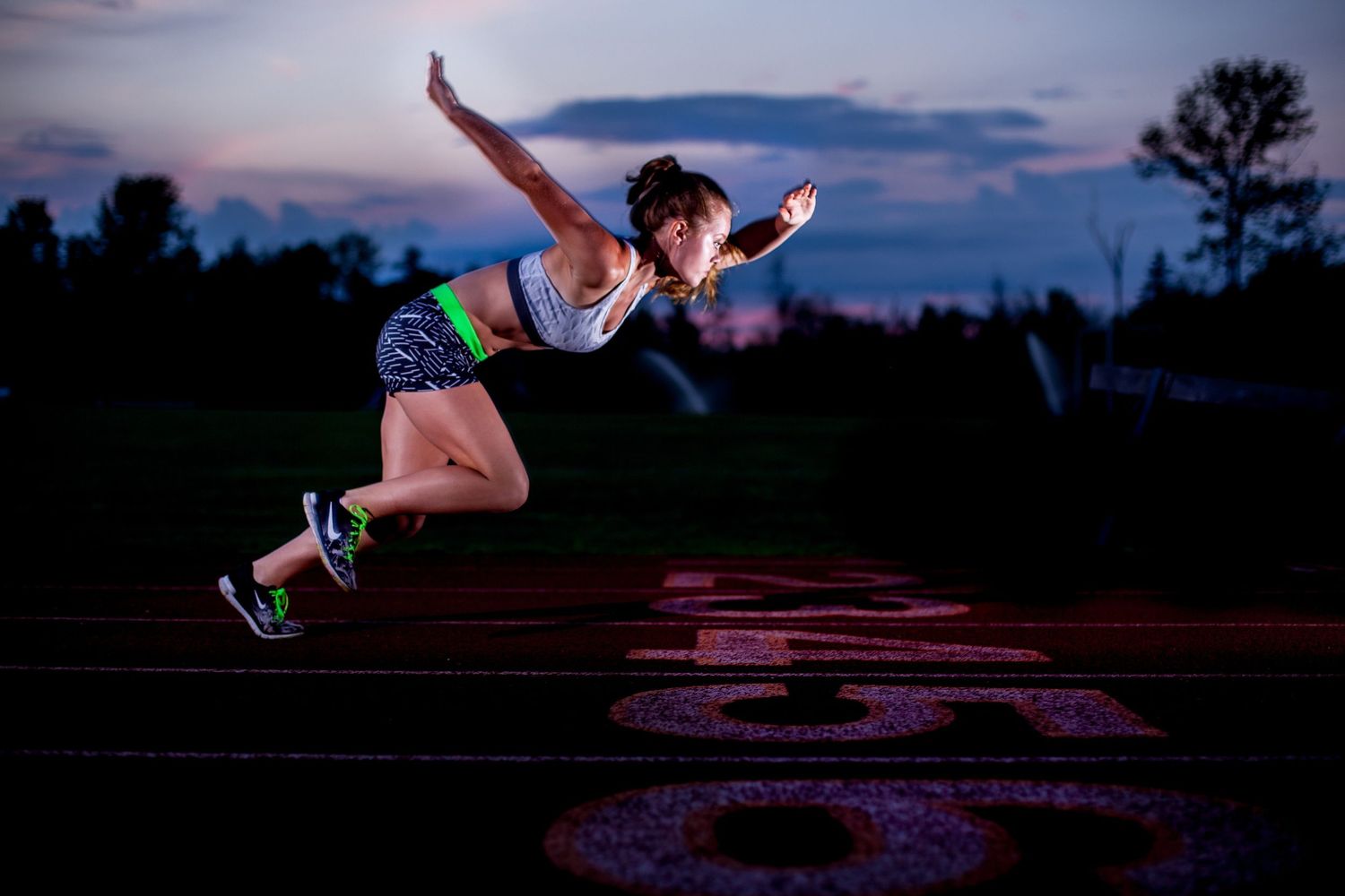 Female athlete captured mid-jump on a running track at twilight under a colorful sky.