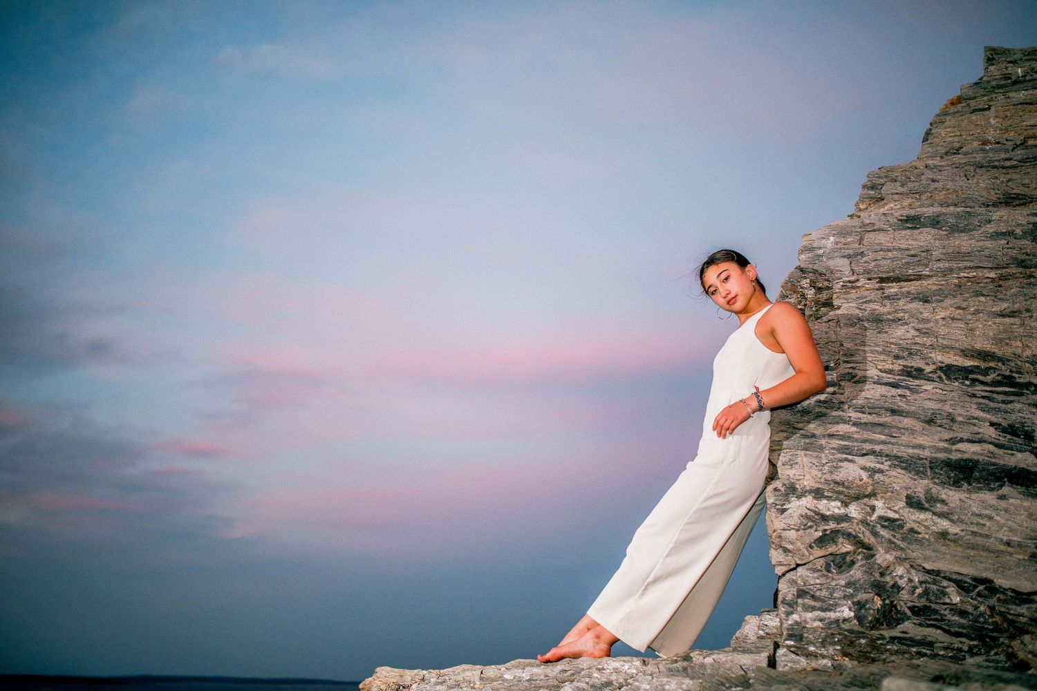 Woman in white jumpsuit leaning against rocky cliff at sunset with ocean in the background.