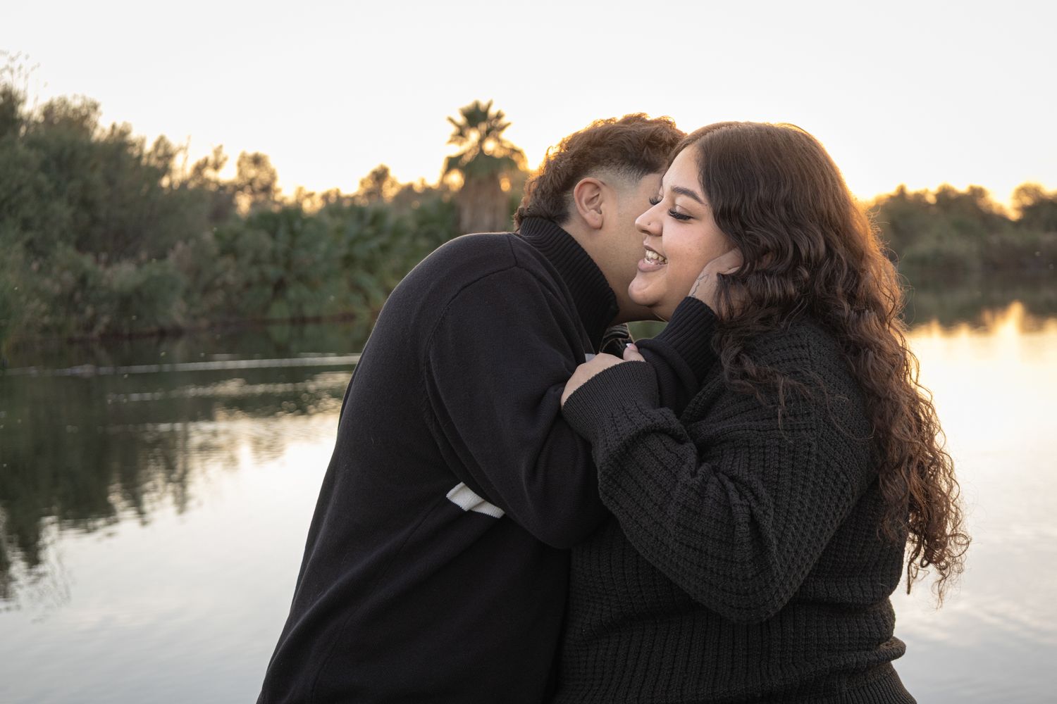 Professional photo, sunset picture over a lake, man cupping a womans face and kissing her cheek, lots of giggles, soft colors