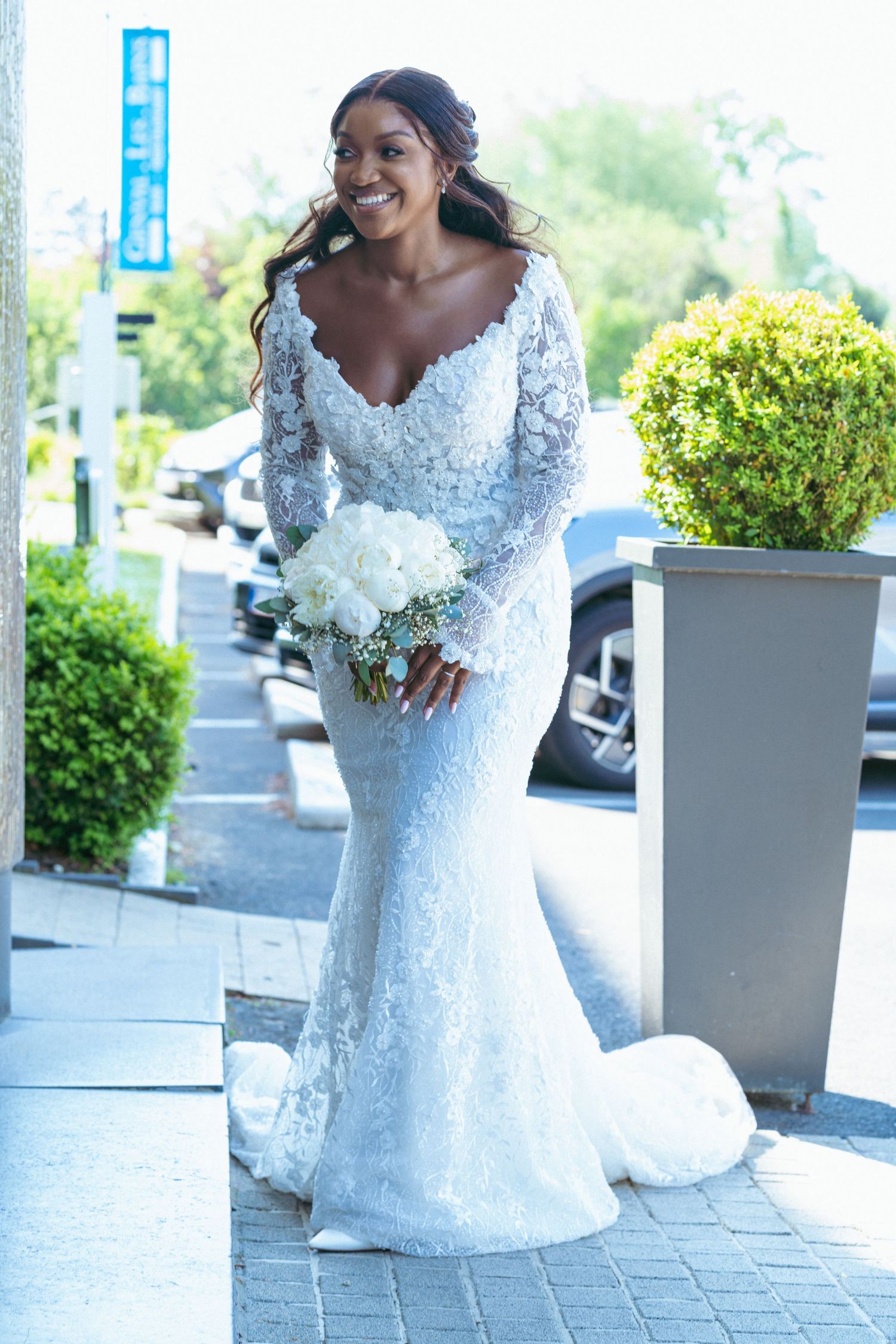 A bride in a lace wedding dress holds a white bouquet while posing outdoors.