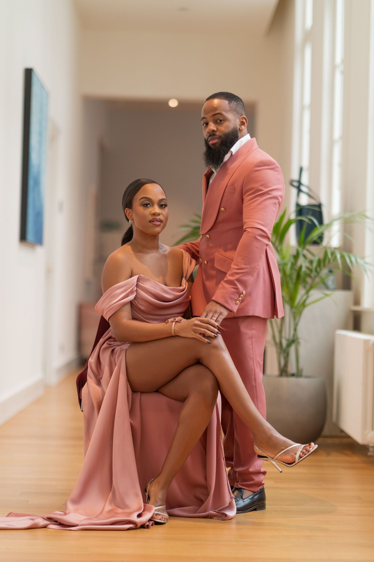 Elegant couple in matching pink formal wear poses in modern hallway setting.