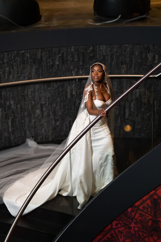 Bride in flowing white gown poses against sleek black car.