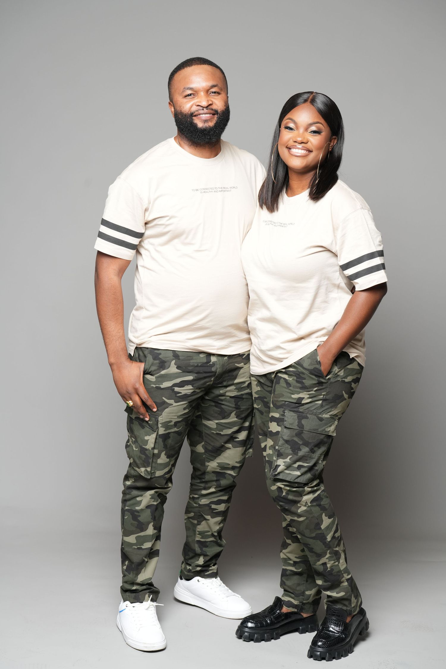 Couple in matching white shirts and camo pants pose against grey backdrop.
