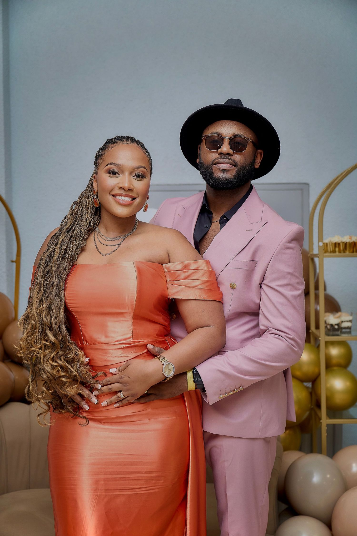 Stylish couple poses in coral dress and pink suit with gold balloon decorations in the background.