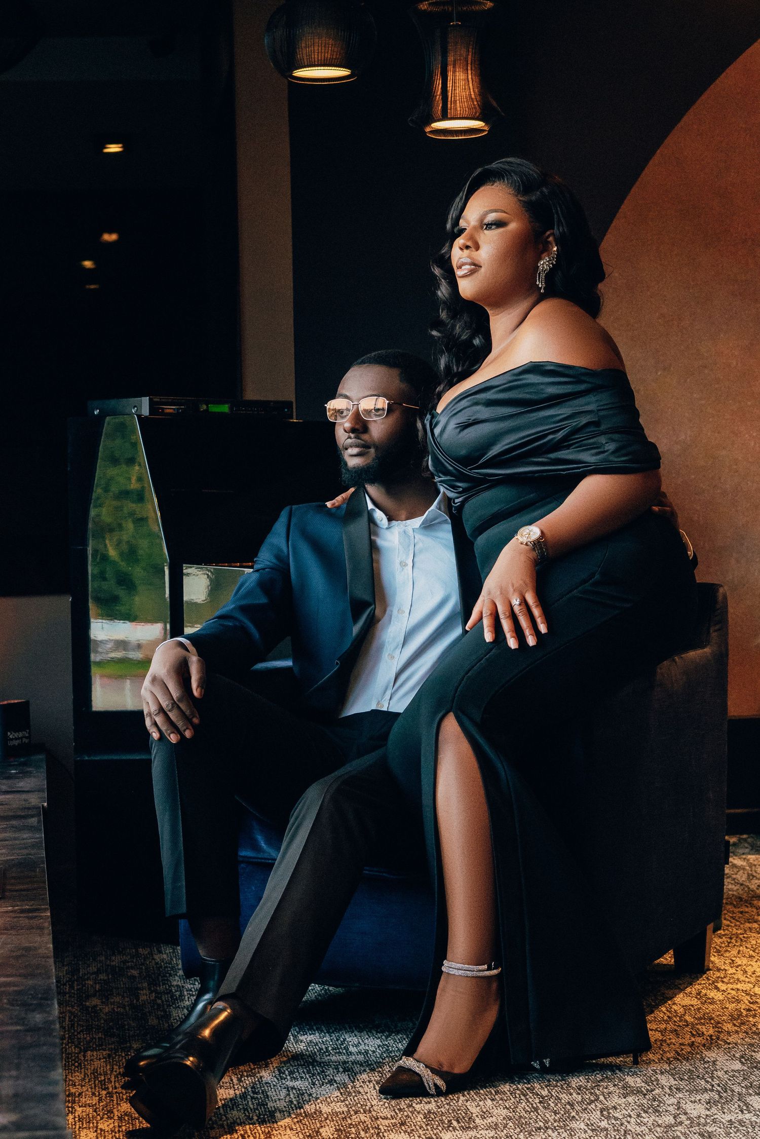Romantic couple in formal attire poses on dark leather couch in moody lighting.