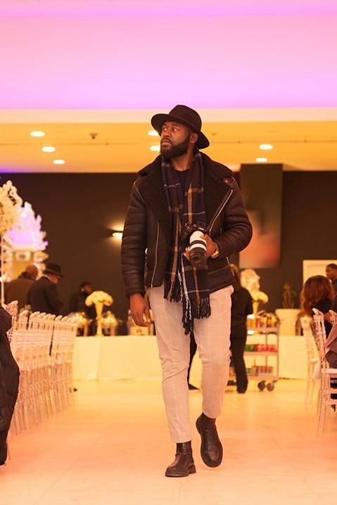 A stylish figure in black attire and hat walks through an elegantly lit indoor space.