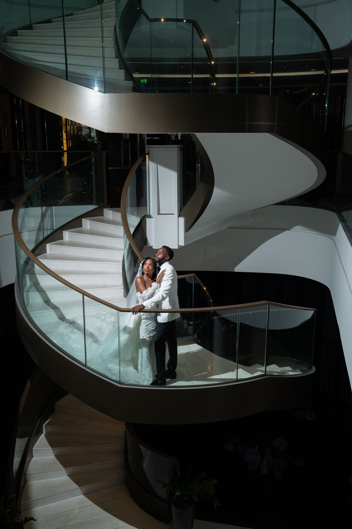 Dramatic wedding photos on modern spiral staircase with glass railings and moody lighting.