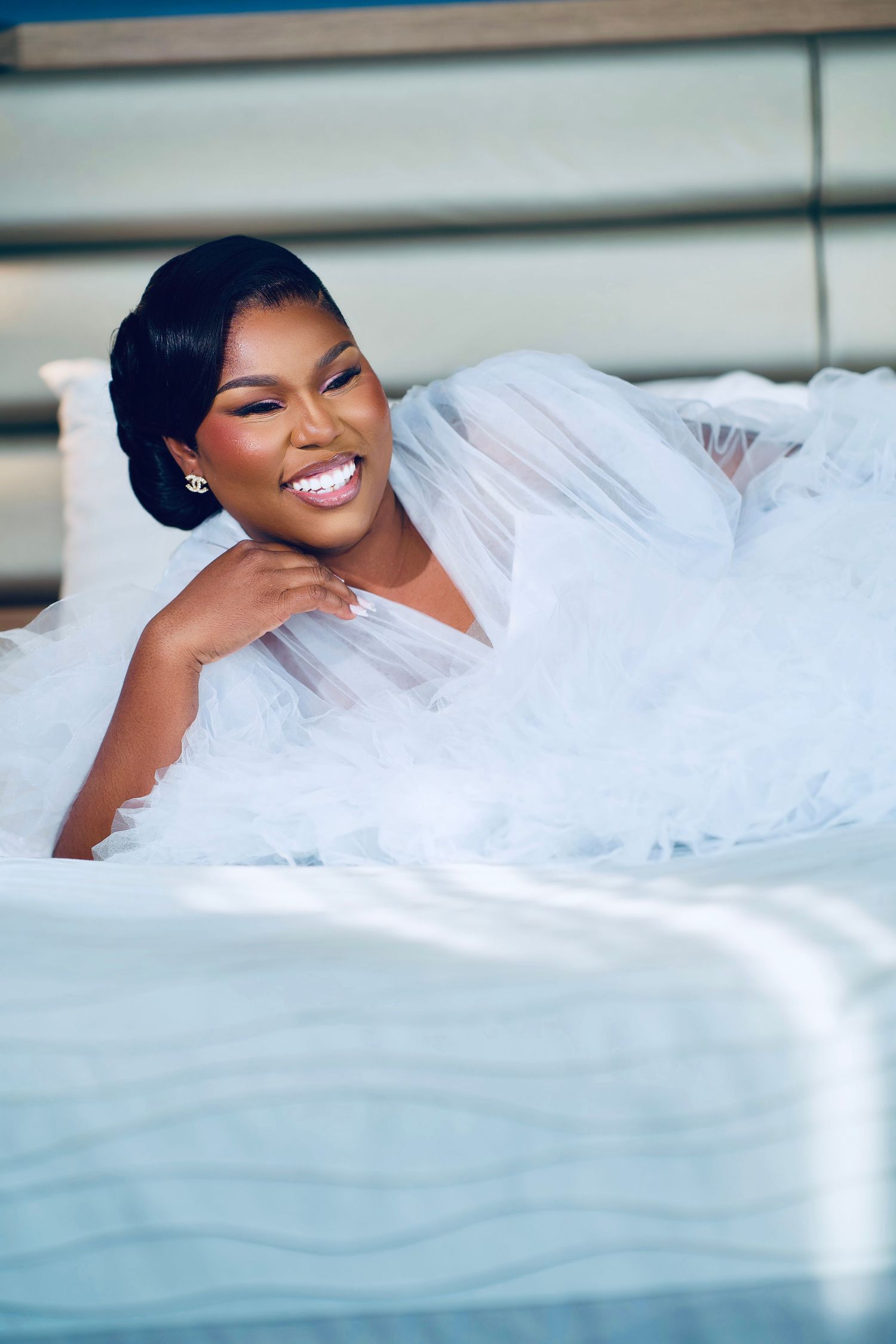 Bride in white dress lounges gracefully on bed while showing genuine joy and laughter.