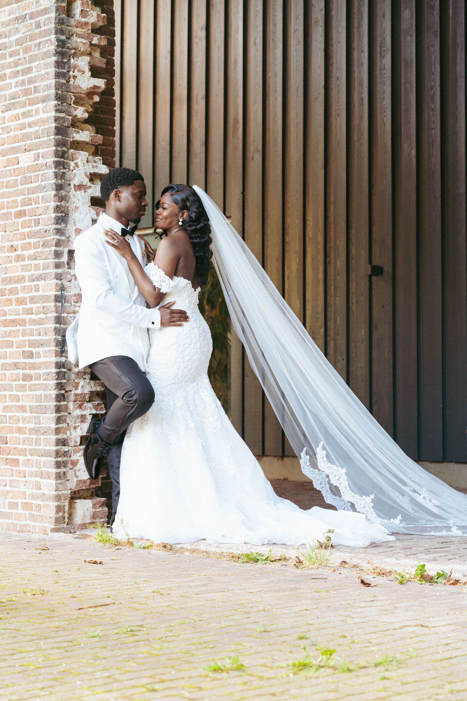 A wedding couple in white attire posing dramatically against a brick and wood architectural backdrop.
