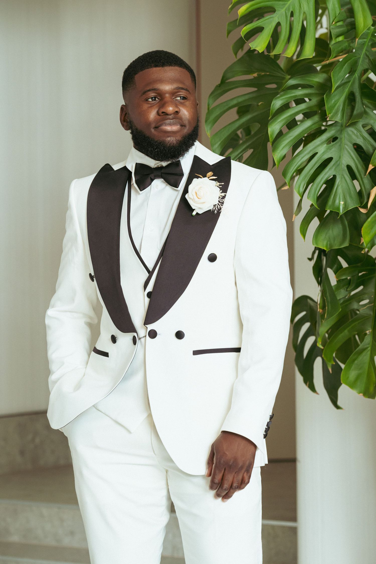 A groom in a stylish white tuxedo with black lapels poses against a backdrop of tropical monstera leaves.
