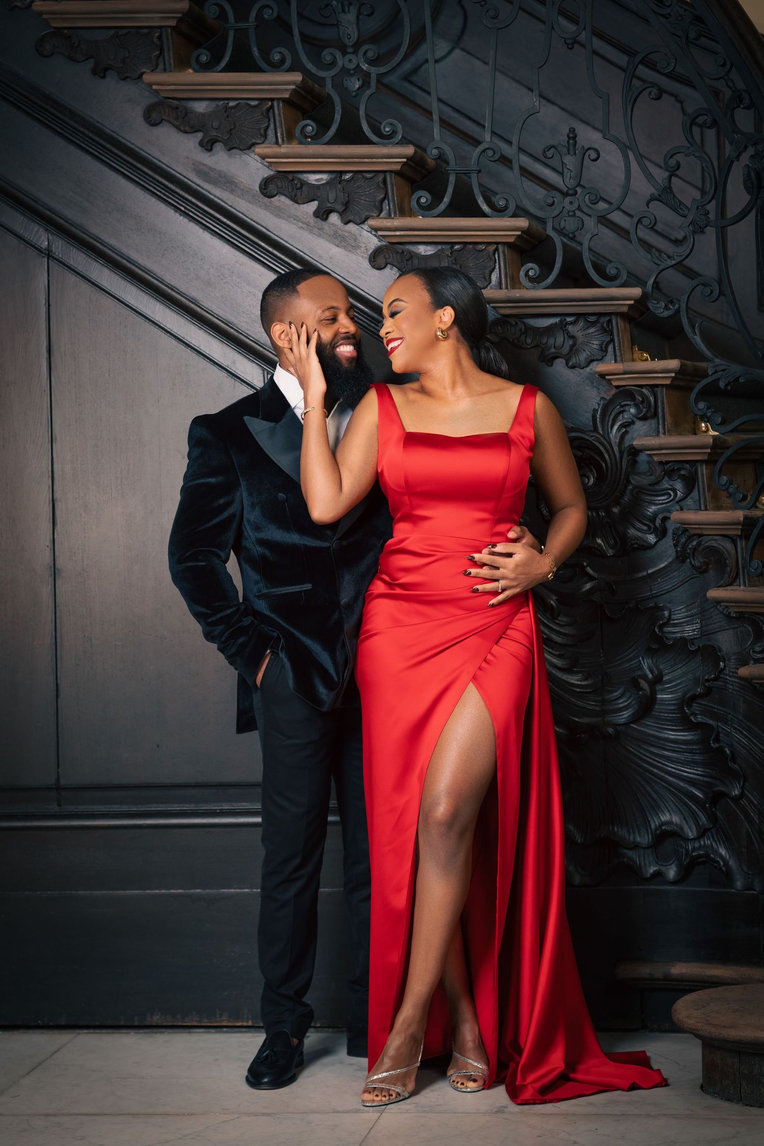 Glamorous couple poses on vintage staircase, wearing black suit and red evening gown.