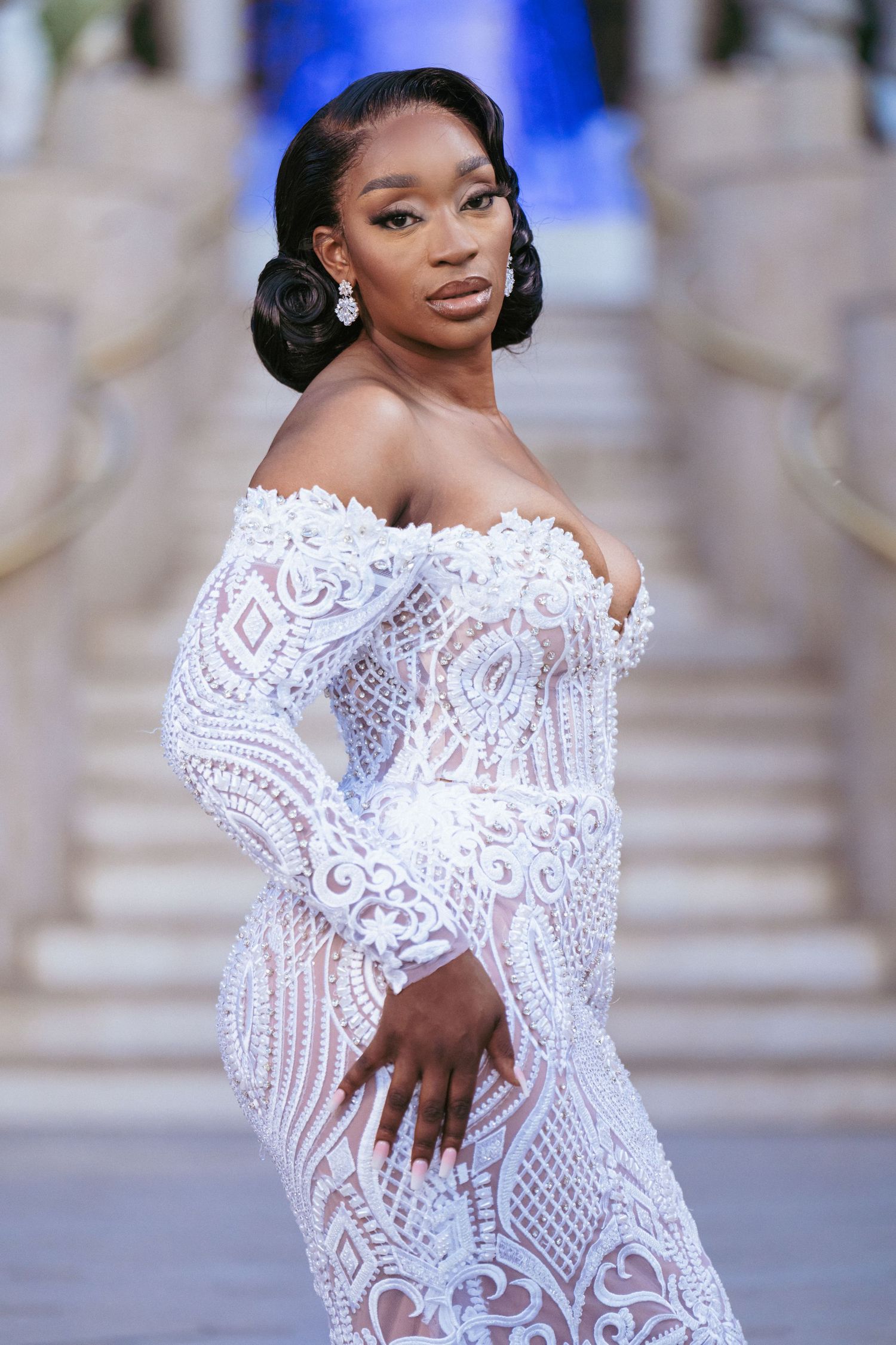 Bride in intricate white lace off-shoulder wedding gown posing elegantly.
