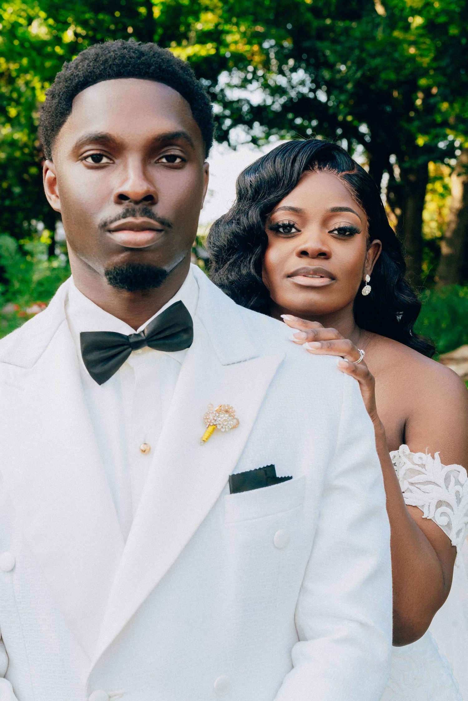 A bride and groom in crisp white formal attire pose together against a natural green background.