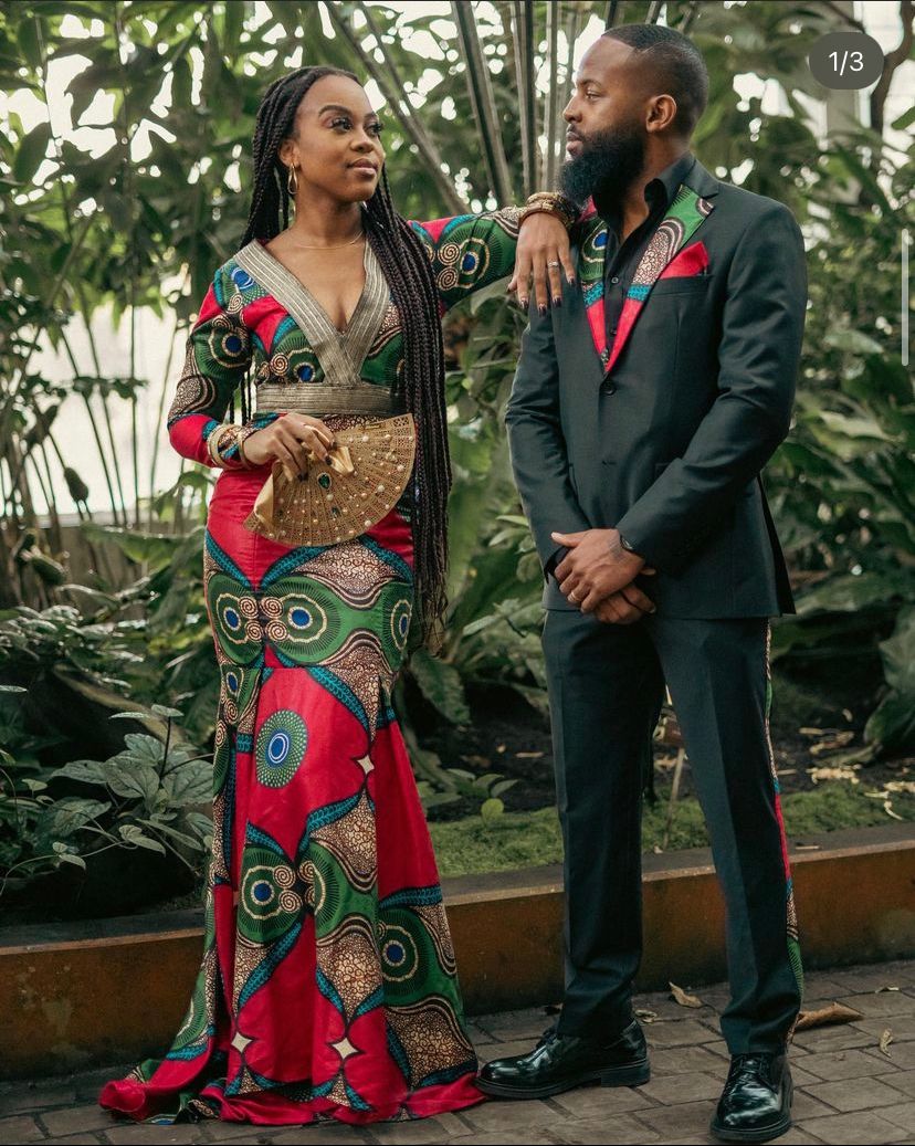 Couple in formal wear poses against tropical backdrop, wearing African print dress and black suit.