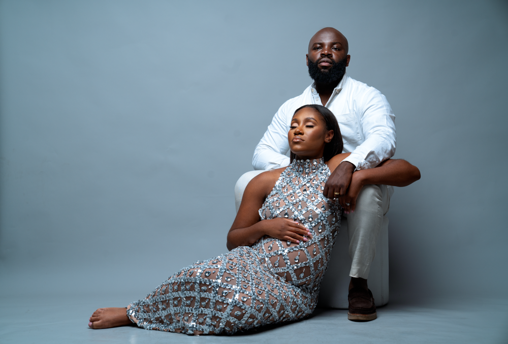 Maternity photoshoot featuring couple in formal wear against blue studio background.