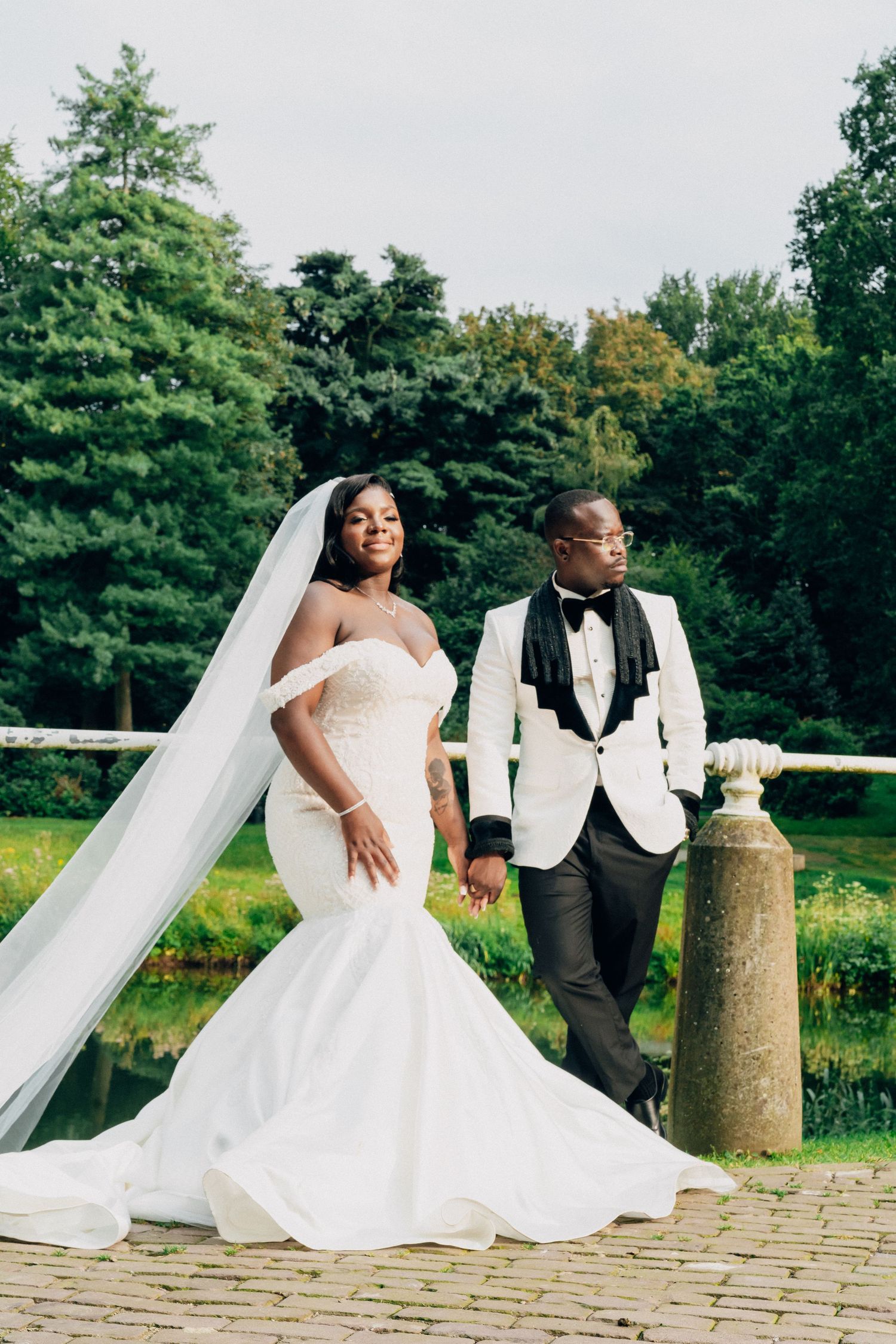 A bride and groom in elegant white attire pose together on stone steps in a garden setting.