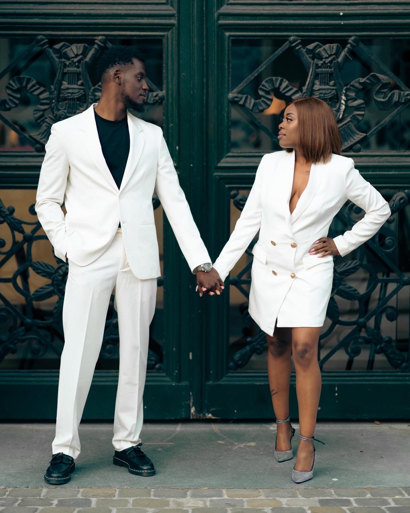 Fashion-forward couple poses in matching white outfits against ornate iron gates.