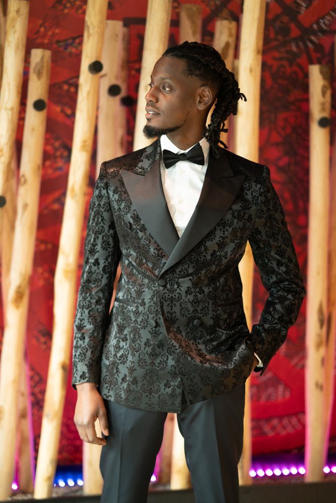 Fashion series showing a black textured jacket against wooden pillars and red backdrop.