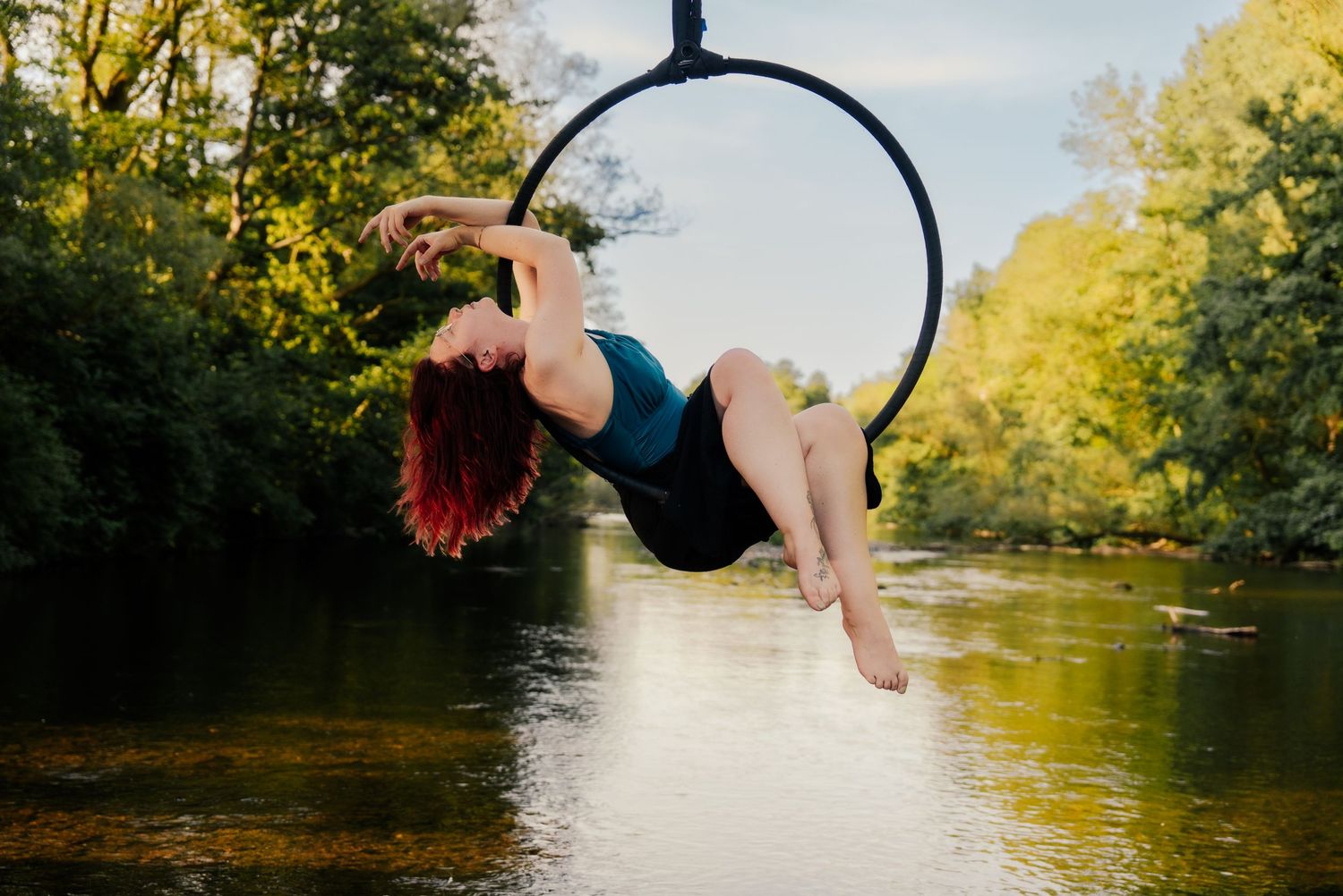 Aerial hoop performer with red hair poses gracefully over a river surrounded by lush green trees.