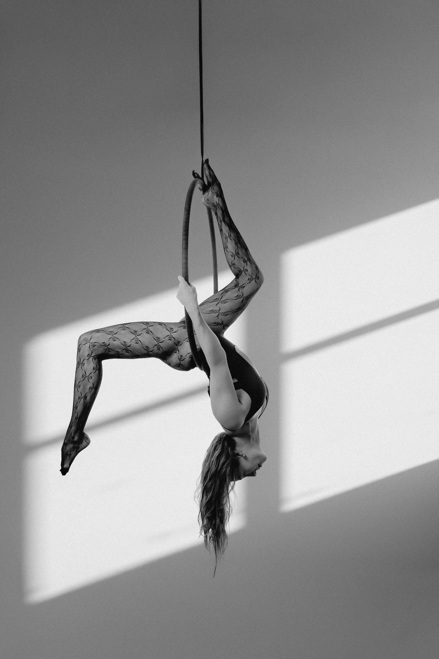 Black and white photo of an aerial acrobat performing an inverted pose on a rope in a lace bodysuit.