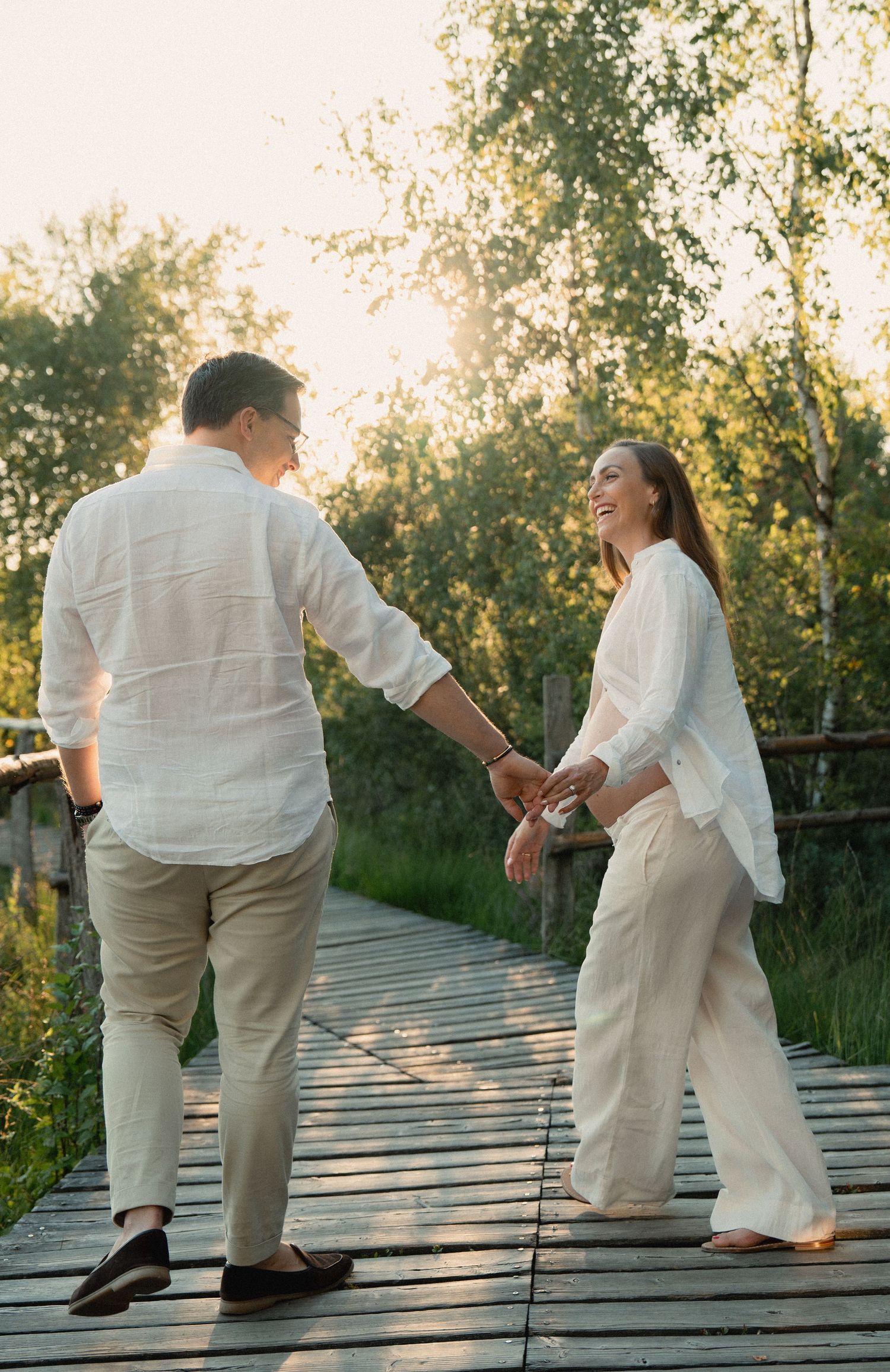 Couple holding hands on a wooden bridge at sunset, dressed in white linen outfits, surrounded by lush green trees.