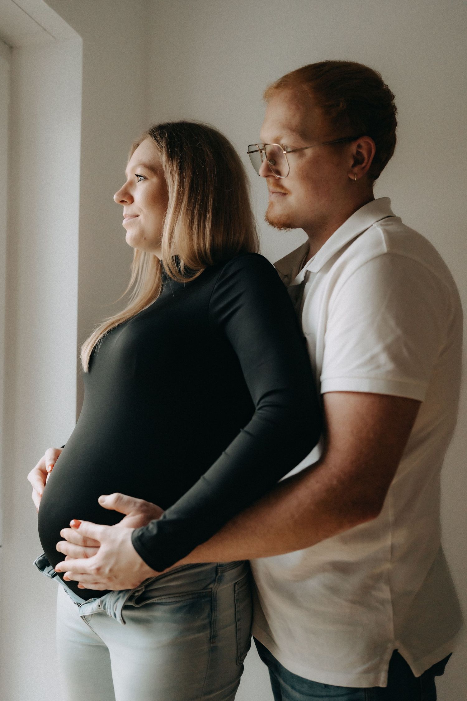 An expecting couple embraces near a bright window in their home.