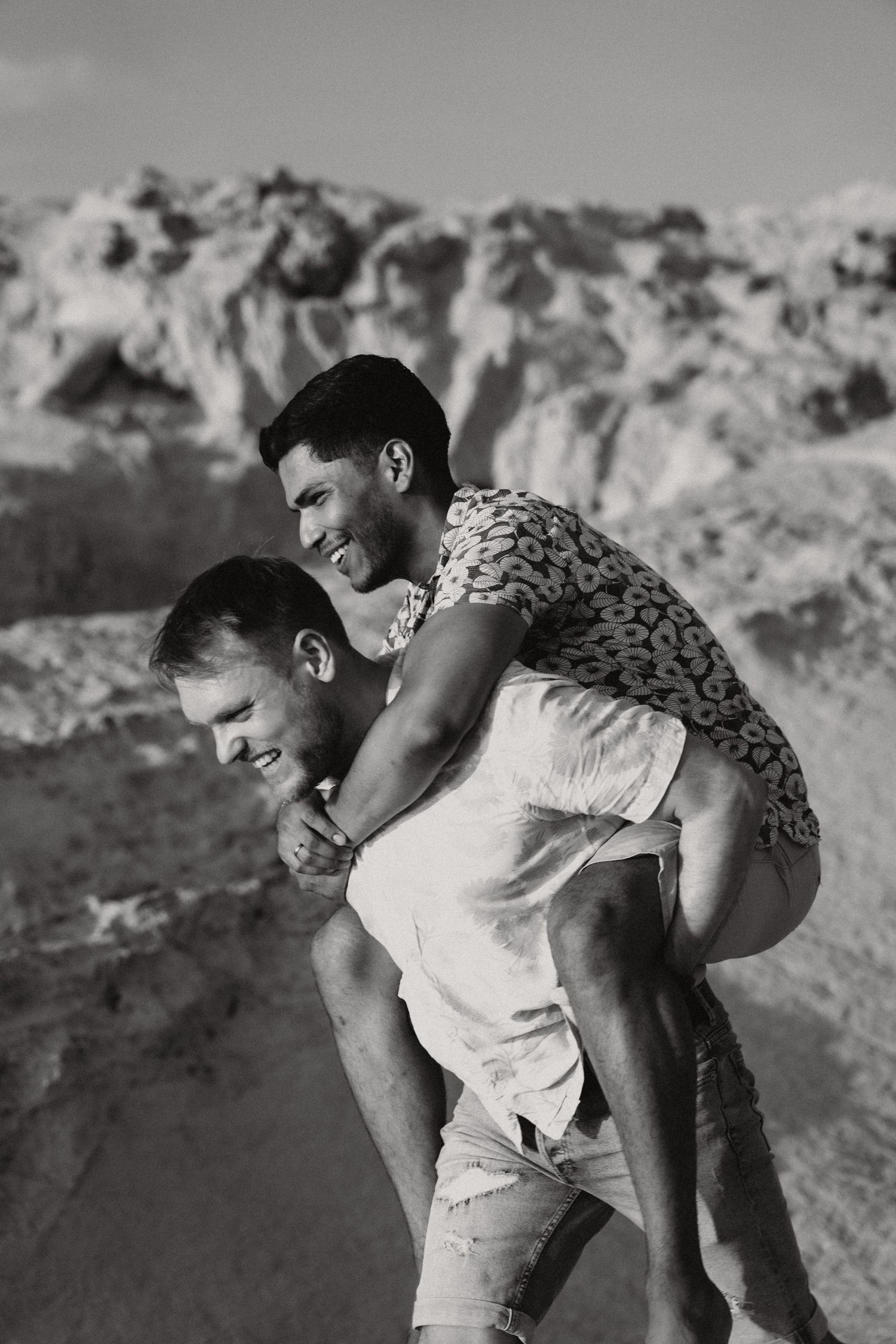 Black and white photo of two young men laughing, one giving the other a piggyback ride on a sandy beach.