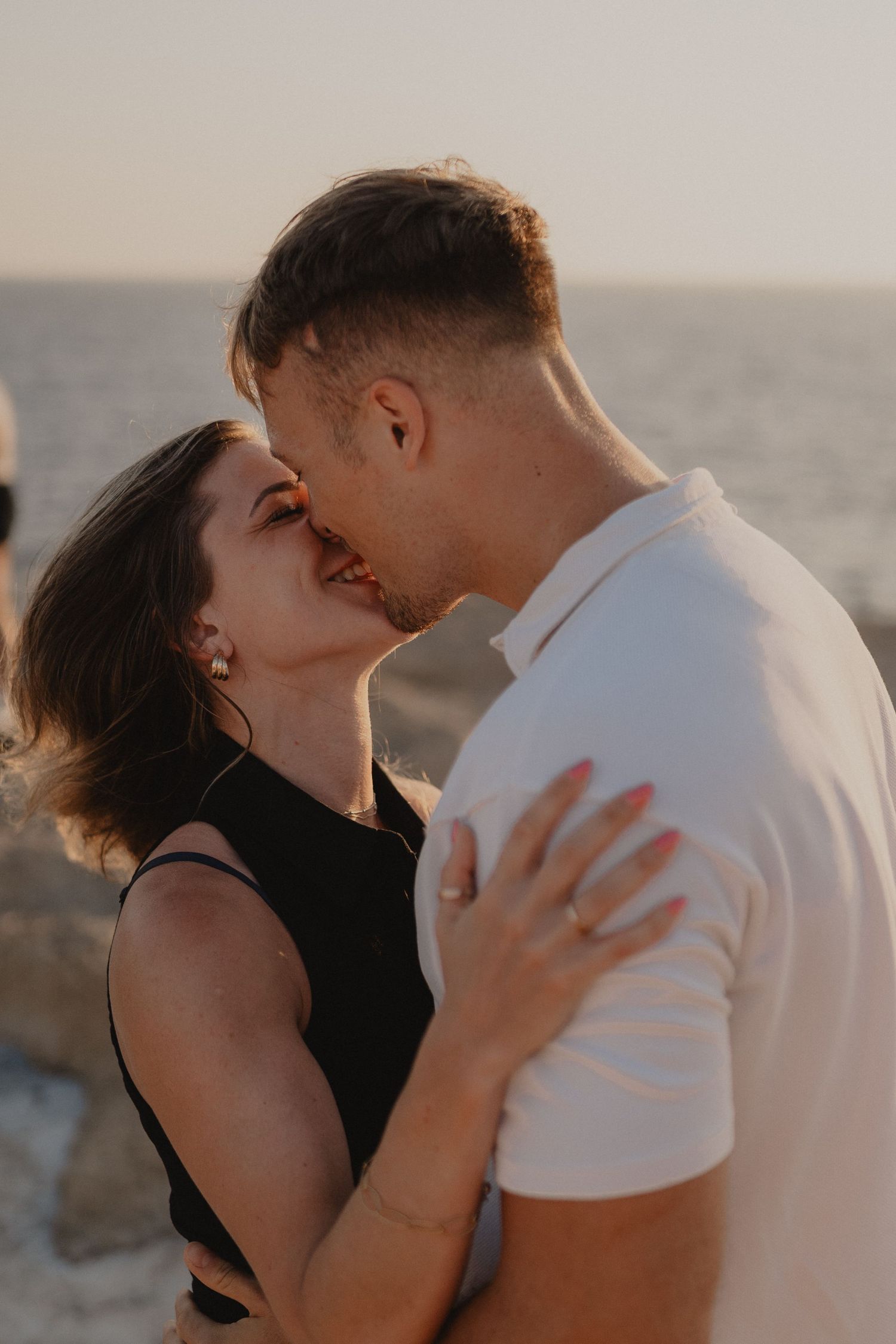 A romantic couple shares a tender kiss by the ocean at sunset, woman in black dress embracing man in white polo shirt.