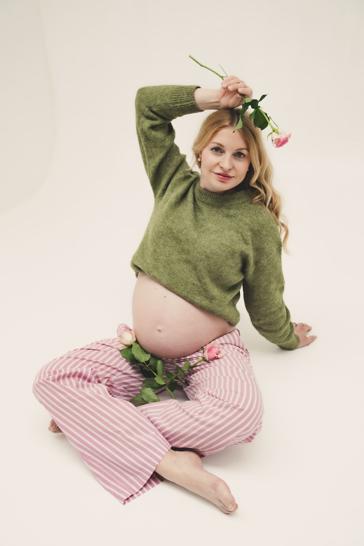 Pregnant woman in green sweater and pink striped pants sits cross-legged holding pink roses, smiling at camera.
