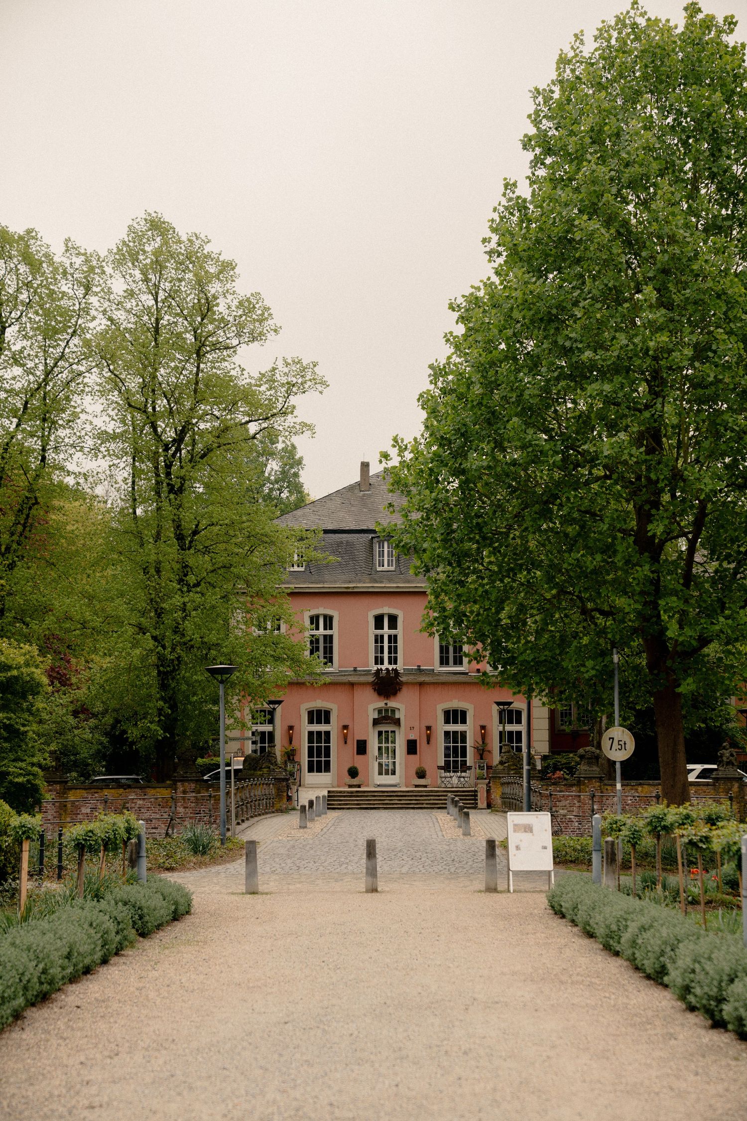 A gravel path leads to a historic pink brick manor house surrounded by lush green trees in a formal garden setting.