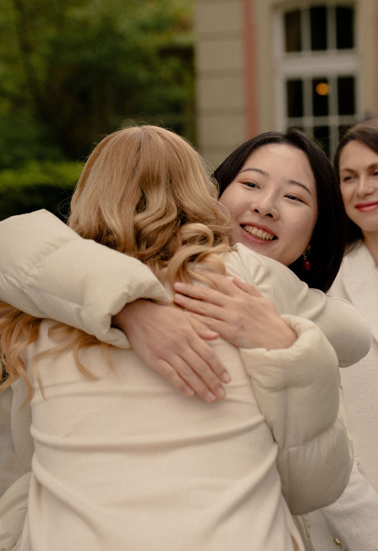 Women in cream puffer jackets sharing warm hugs outdoors at an elegant event.