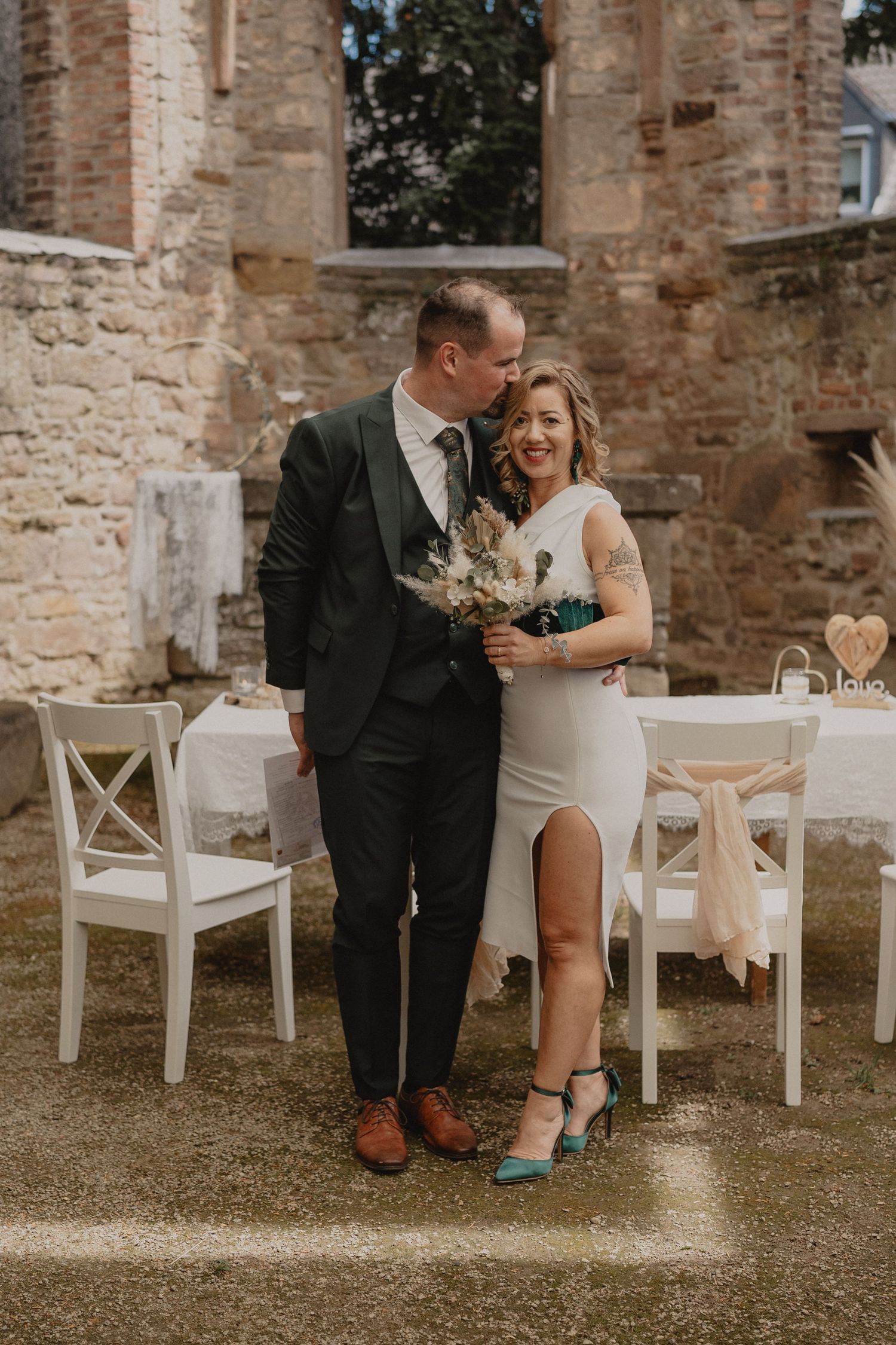 Couple shares an intimate moment on white chairs against a historic stone wall background.